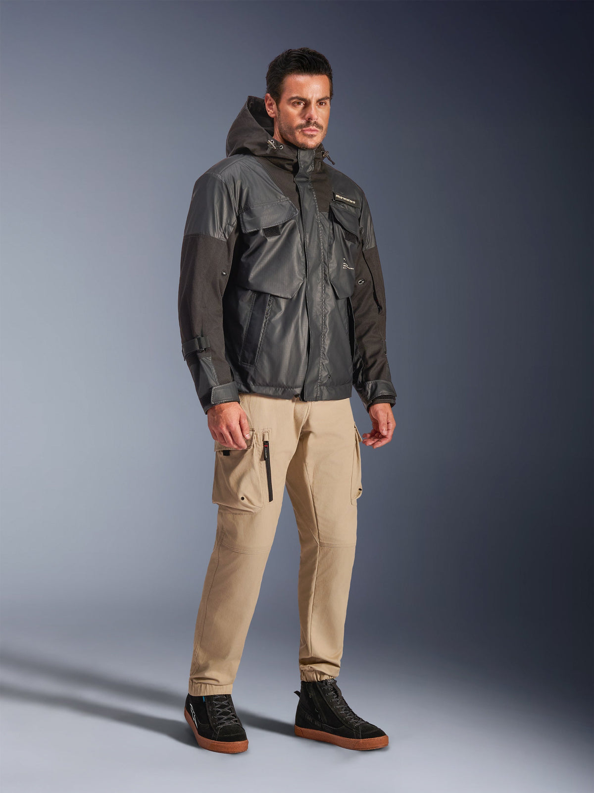 Modelo masculino a usar o Casaco Impermeável Alpinestars Mospher em preto, parka impermeável de inspiração utilitária com design multibolsos e capuz, calças cargo bege e sapatilhas pretas de cano alto, equipamento urbano para motociclistas