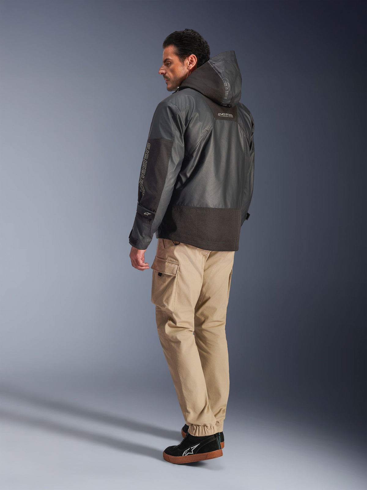 Homem a usar o Casaco Mospher WP da Alpinestars em preto, vista de perfil lateral e traseira, combinado com calças cargo bege e ténis pretos. Apresenta um design de parka com capuz em tecido ripstop, logótipo Alpinestars na manga e um estilo de motociclismo urbano moderno.