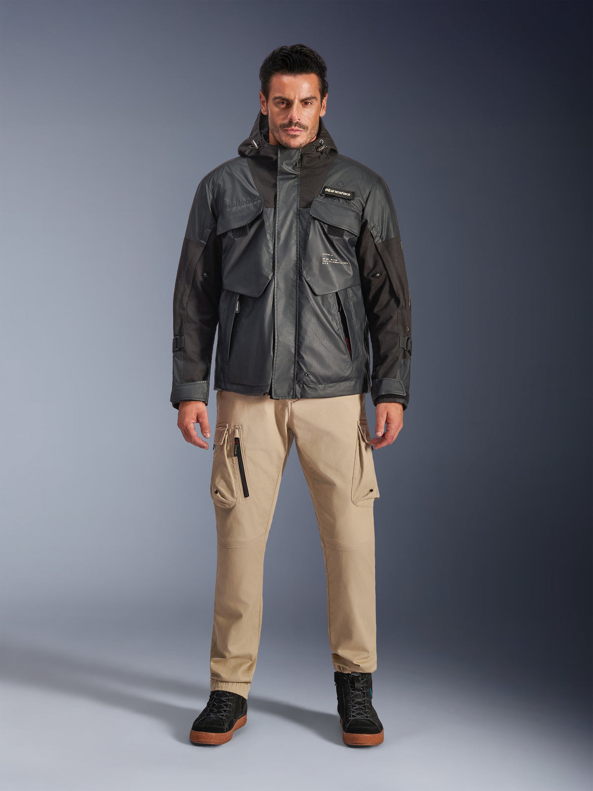 Modelo masculino a usar o Casaco Alpinestars Mospher Waterproof em preto, equipamento de motociclismo urbano contemporâneo, design tático utilitário com múltiplos bolsos funcionais e capuz, combinado com calças cargo bege e sapatos pretos, retrato de corpo inteiro em estúdio.