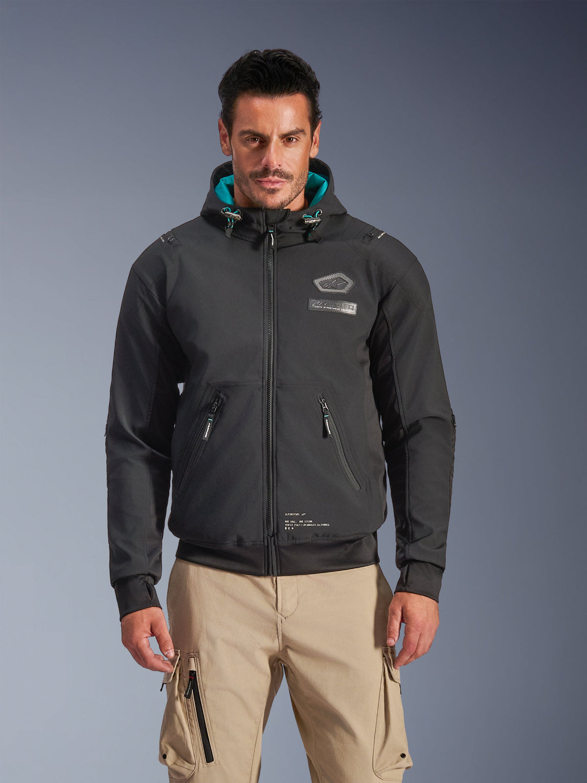 Alpinestars Moblast Waterproof Jacket, Men's Urban Motorcycle Jacket, Black with Teal Hood Lining, features 3D zip pockets and hooded design, worn with tan cargo pants, modern urban motorcycle apparel