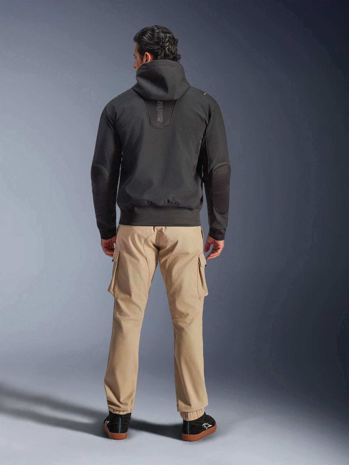 Alpinestars Moblast Waterproof Jacket, Black, back view of urban motorcycle hoodie, worn with tan cargo pants and black sneakers, minimalist design with vertical 'astars' branding on the upper back, contemporary all-weather riding gear