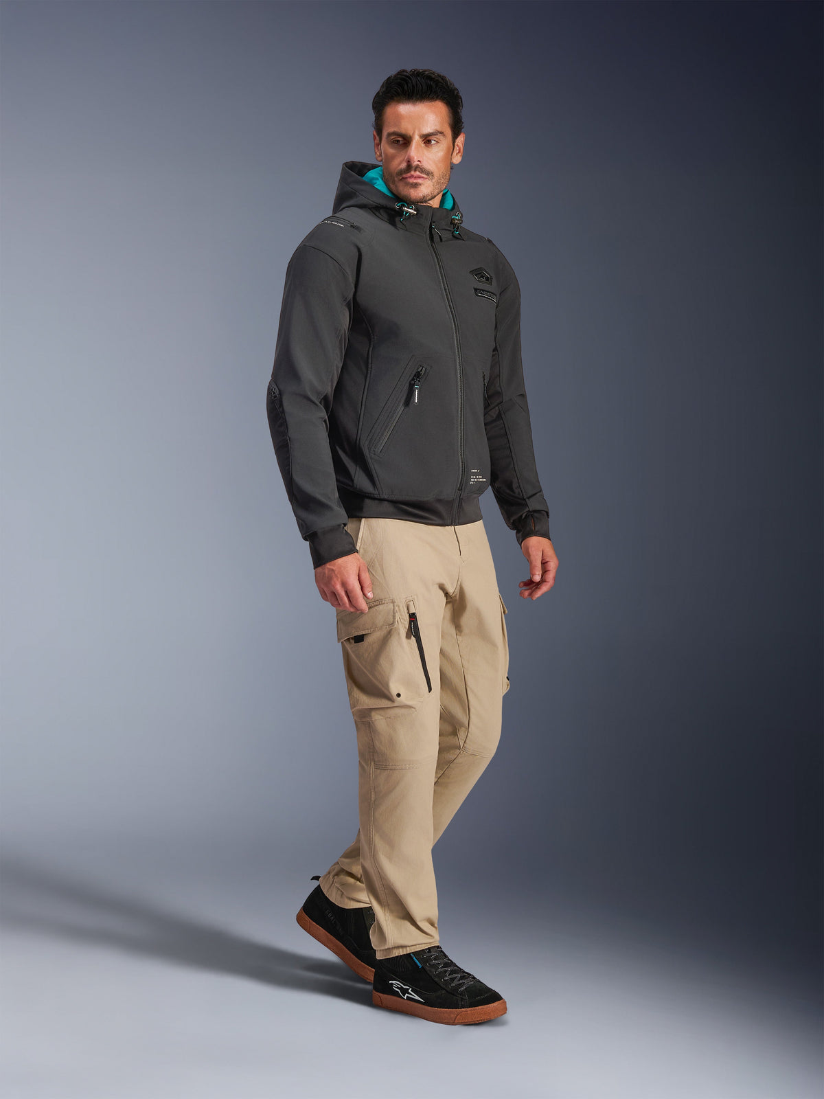 Male model wearing Alpinestars Moblast Waterproof Jacket in Black, urban motorcycle hoodie with teal-lined hood, paired with khaki cargo pants and black sneakers, modern minimalist design with technical stretch fabric for all-weather commuting