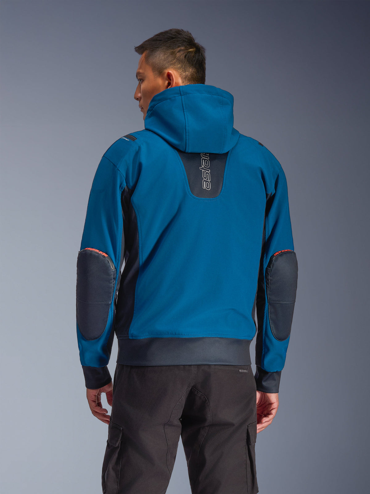 Alpinestars Moblast Waterproof Jacket, em Dark Petrol e Dark Navy, hoodie de motociclismo urbano visto de costas, com capuz fixo, logótipo 'astars' vertical na parte superior das costas, remendos reforçados escuros nos cotovelos, cintura e punhos elásticos, vestuário técnico de condução moderno