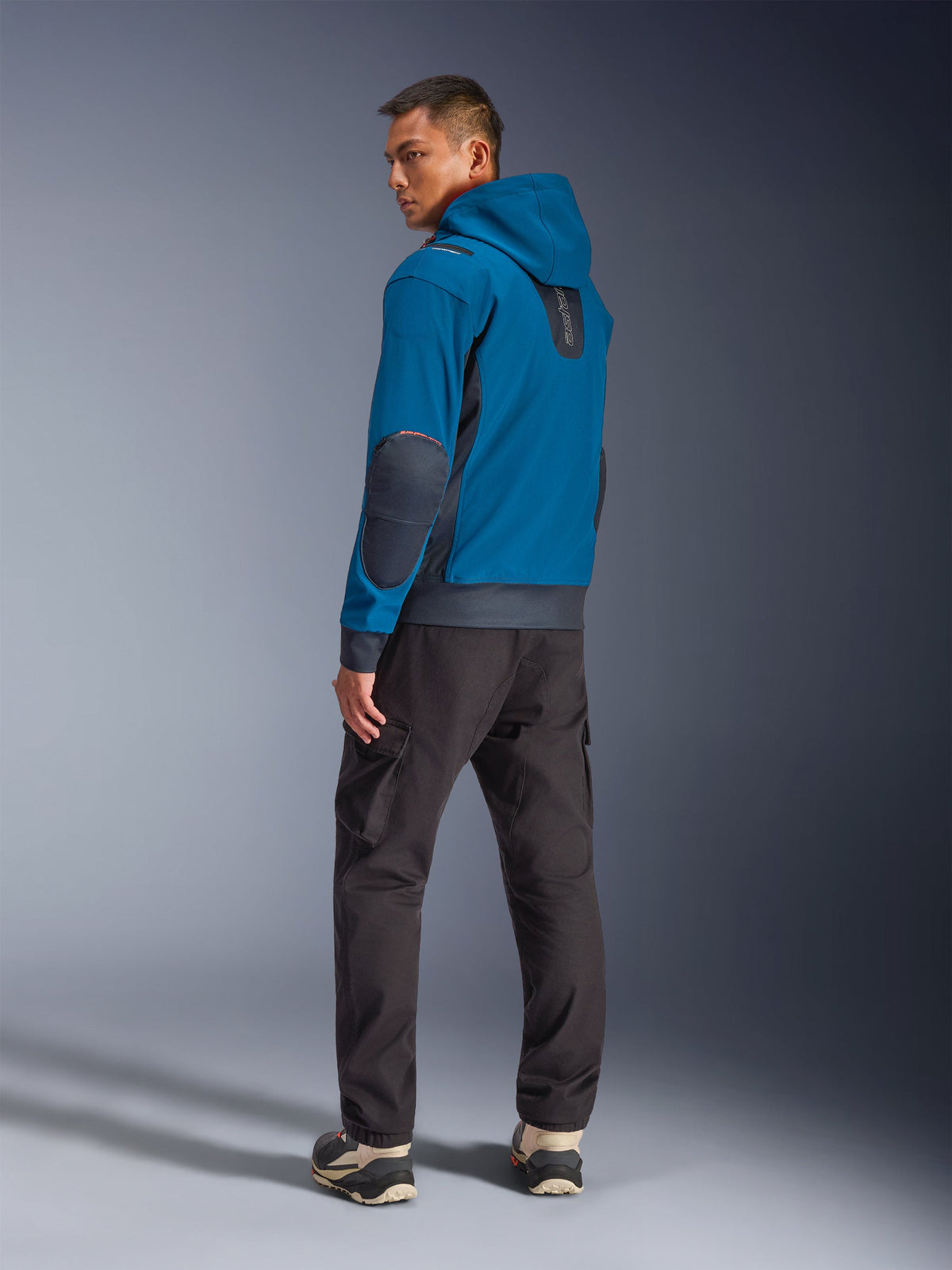 Casaco Impermeável Alpinestars Moblast em Dark Petrol e Dark Navy, vista traseira num modelo mostrando o design com capuz, remendos nos cotovelos e inserções laterais em azul-marinho escuro, combinado com calças cargo escuras e sapatilhas técnicas, vestuário de motociclismo urbano moderno.