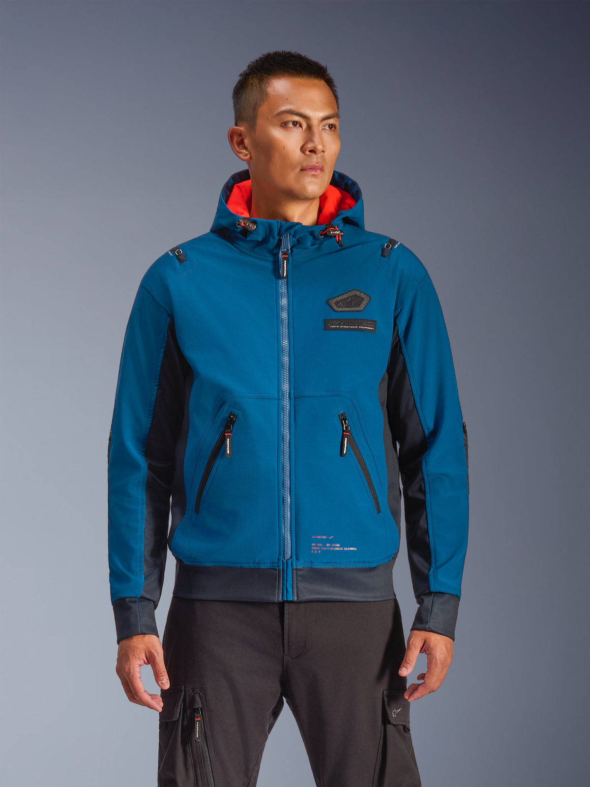 Casaco Alpinestars Moblast Waterproof usado por modelo, azul e azul marinho escuro, casaco de motociclismo urbano, design com capuz e forro interior vermelho, fechos de correr pretos e detalhe de emblema no peito, equipamento de motociclismo inspirado em streetwear