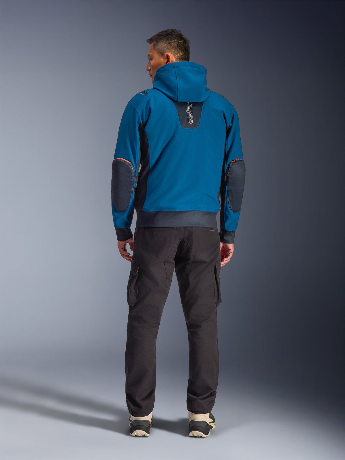 Casaco Alpinestars Moblast Waterproof, casaco de motociclismo urbano, Dark Petrol e azul marinho escuro, vista traseira mostrando o capuz e reforços nos cotovelos, usado com calças cargo escuras, equipamento de condução elegante para todas as condições climatéricas