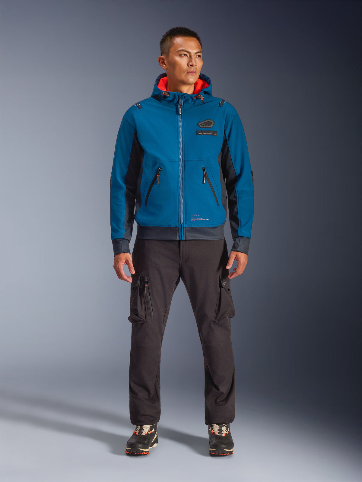 Casaco Impermeável Alpinestars Moblast, casaco de motociclismo com capuz em azul Dark Petrol e Dark Navy com forro interior cor-de-laranja, vestido por um modelo masculino com calças cargo escuras e sapatilhas pretas, equipamento de condução urbana moderno