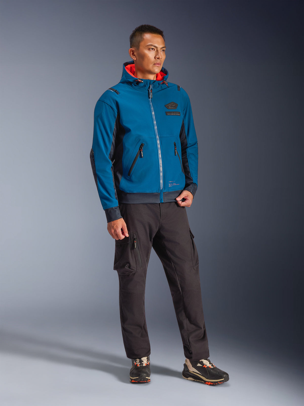 Modelo masculino a usar o casaco impermeável Alpinestars Moblast nas cores Dark Petrol e Dark Navy, com design de capuz e interior em laranja vibrante, combinado com calças cargo pretas e ténis, equipamento de motociclismo urbano moderno.