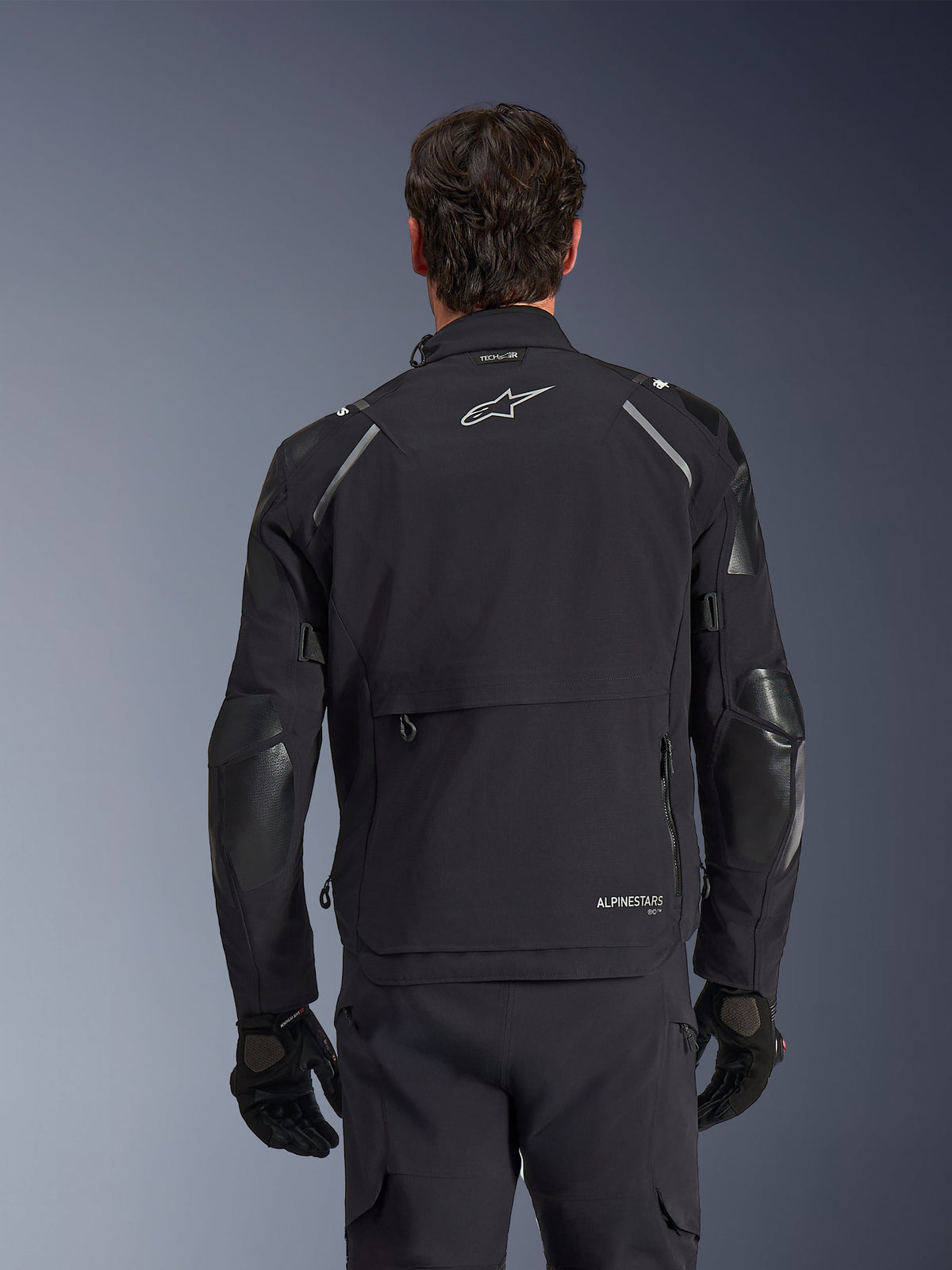 Casaco Alpinestars Andes Pro Drystar® XF, preto e cinzento escuro, vista traseira num modelo masculino, com um compartimento de armazenamento traseiro grande, detalhes refletores nos ombros e logótipo da marca, combinado com luvas e calças pretas para motociclismo de aventura.