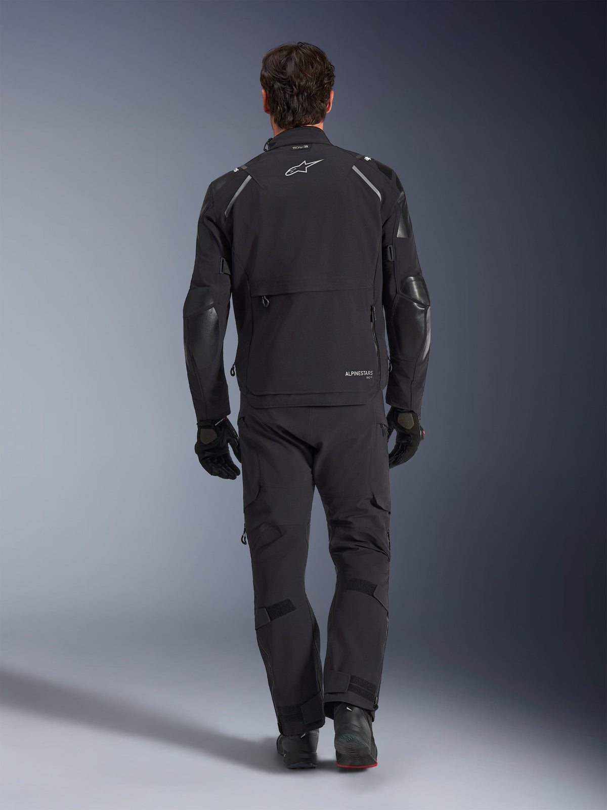 Casaco Alpinestars Andes Pro Drystar® XF, casaco de turismo de aventura, preto e cinzento escuro, vista traseira com grande bolso de carga e painéis reforçados nos cotovelos, usado com calças e luvas pretas a condizer, design funcional para condução em todas as condições meteorológicas