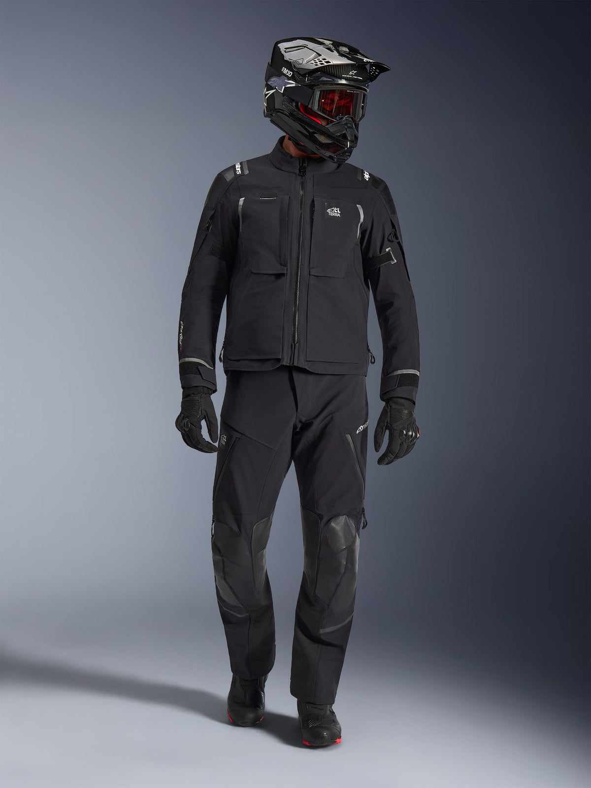 Motociclista de aventura a usar o casaco Alpinestars Andes Pro Drystar® XF em preto e cinzento escuro, calças de aventura pretas a condizer, capacete preto e branco com óculos vermelhos, luvas e botas pretas; apresenta grandes bolsos utilitários frontais, detalhes refletores e construção técnica laminada de 3 camadas para touring em todas as condições climáticas.