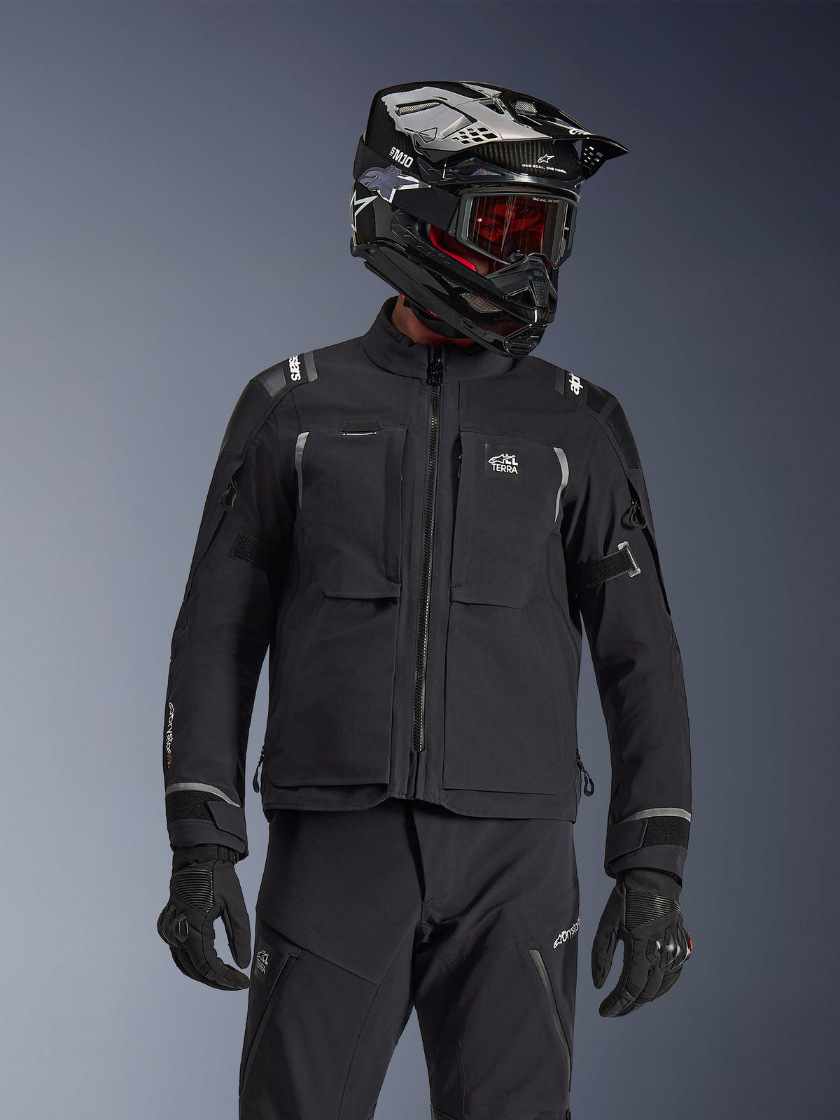 Motociclista de aventura a usar o Casaco Alpinestars Andes Pro Drystar® XF em preto e cinzento escuro, com a marca All Terra no peito, combinado com calças técnicas a condizer e um capacete de aventura preto com óculos, conjunto de equipamento profissional de touring.