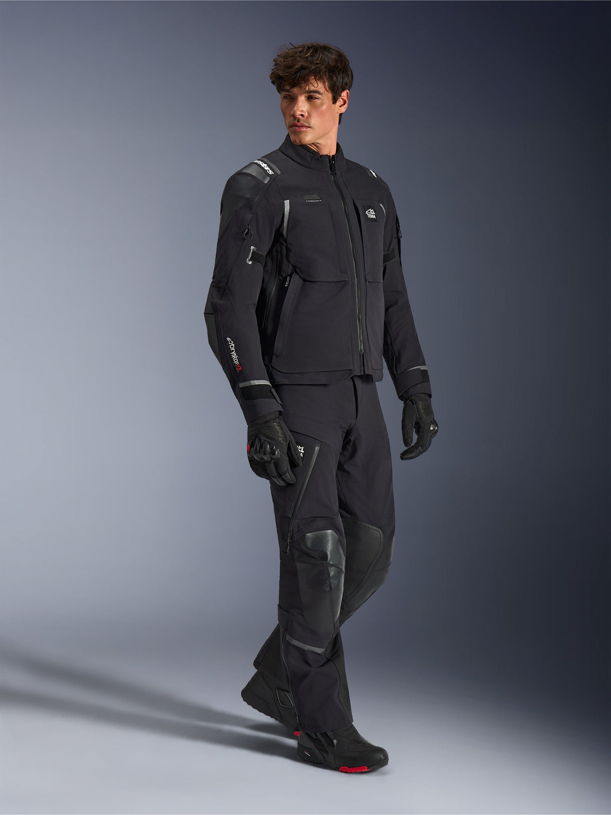 Casaco Alpinestars Andes Pro Drystar® XF, casaco de motociclismo preto e cinzento escuro, vestido por modelo masculino com calças de aventura, luvas e botas a condizer, fotografia de estúdio de corpo inteiro, equipamento técnico de touring e aventura com detalhes refletores e logótipos da marca