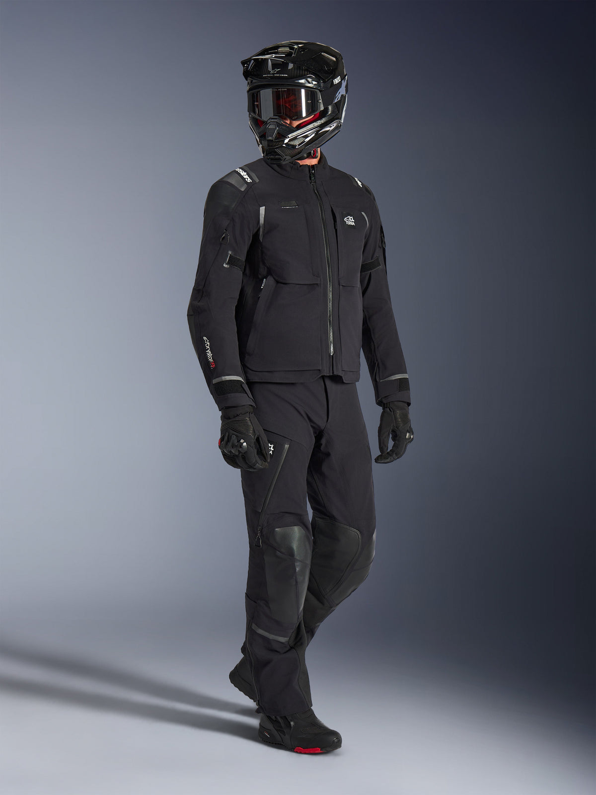Casaco Alpinestars Andes Pro Drystar® XF, casaco de aventura para motociclismo em preto e cinzento escuro, usado com calças têxteis pretas a condizer, capacete integral preto com óculos, luvas e botas, apresentando um revestimento laminado de 3 camadas e múltiplos bolsos de armazenamento para condução de aventura em todas as estações.