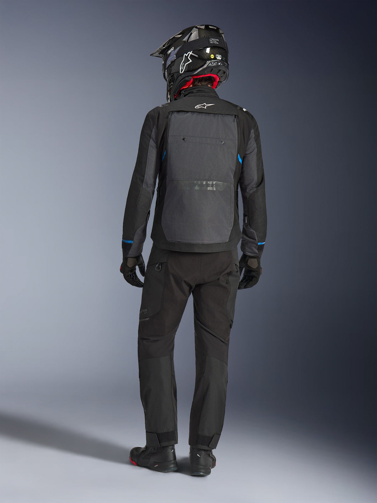 Casaco Alpinestars Cusco Drystar®, preto e cinzento escuro, vista traseira de um condutor com equipamento completo de touring de aventura, grande painel traseiro cinzento com o logótipo Alpinestars, detalhes azuis nas mangas, usado com calças pretas a condizer e capacete.