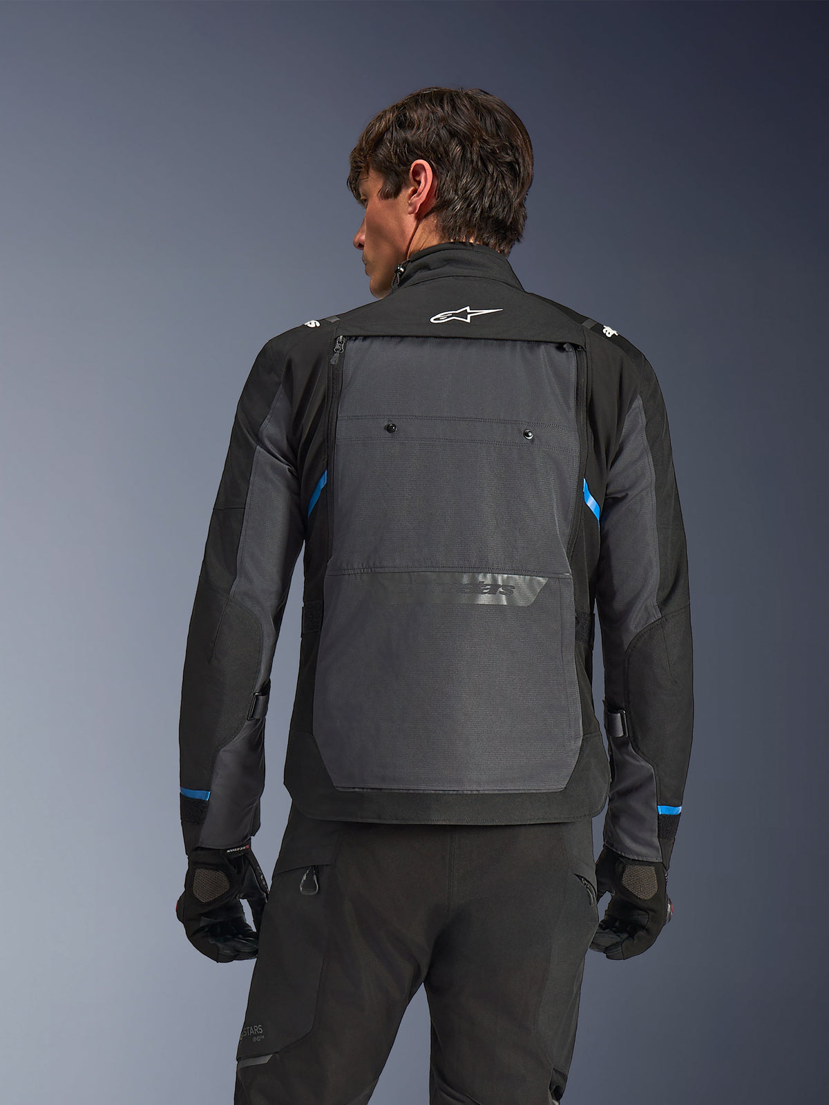 Casaco Alpinestars Cusco Drystar®, casaco de motociclismo, preto e cinzento escuro, vista traseira num modelo mostrando um grande painel traseiro cinzento, logótipo Alpinestars branco e detalhes azuis nas mangas.
