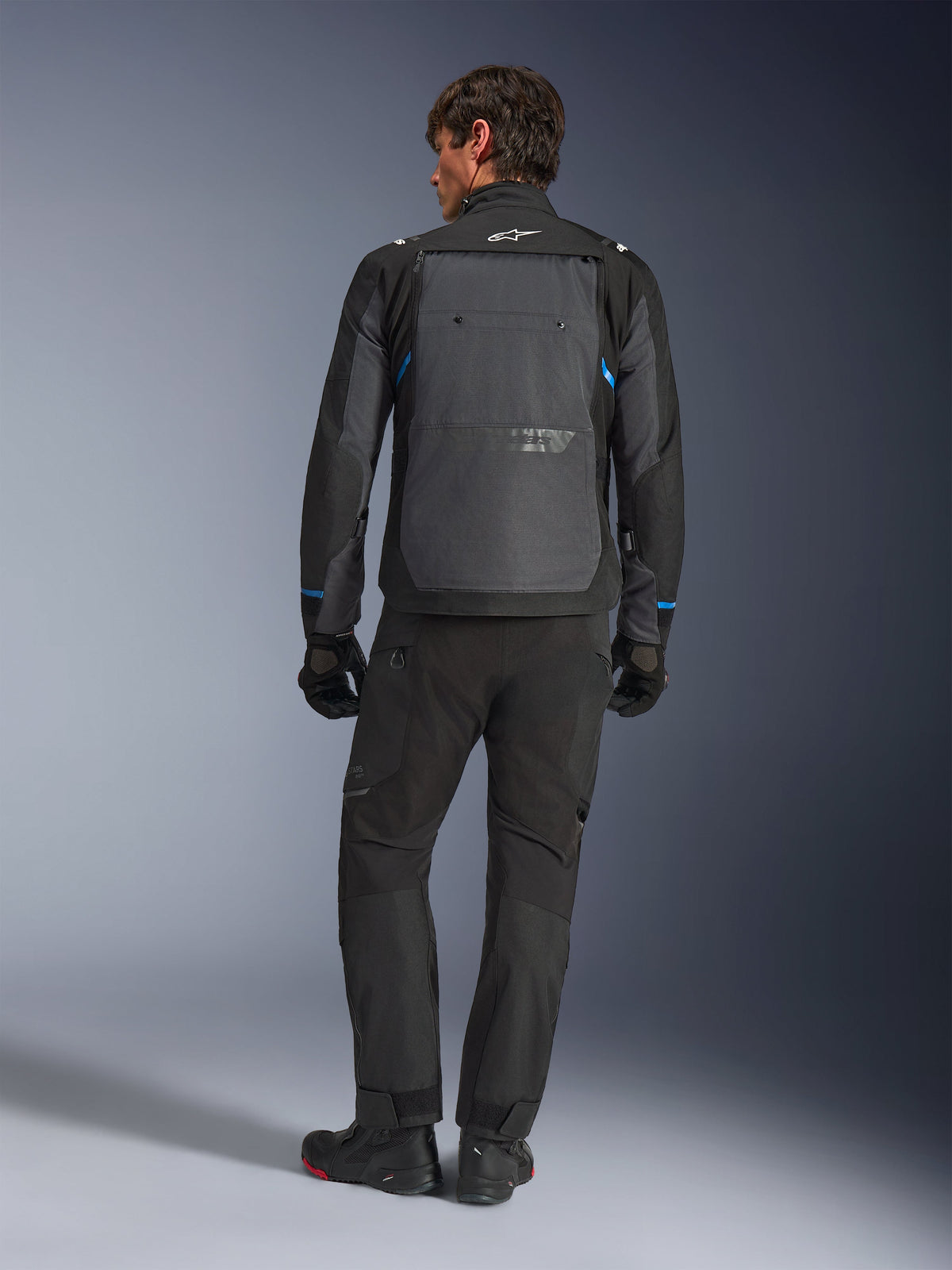 Casaco Alpinestars Cusco Drystar®, preto e cinzento escuro com detalhes azuis, vista traseira, usado com calças de aventura e luvas pretas a condizer, com um grande painel traseiro cinzento e logótipo branco na gola, concebido para estrada e turismo de aventura