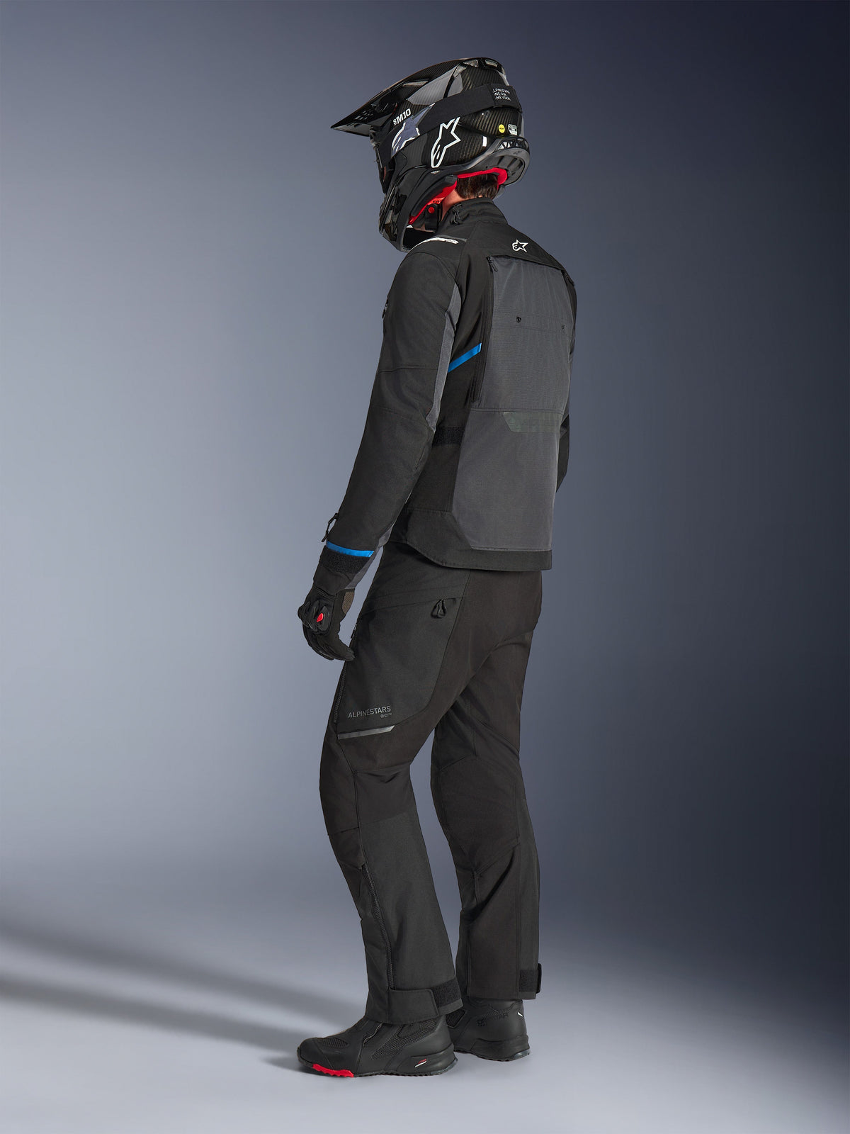 Casaco Alpinestars Cusco Drystar® em preto e cinzento escuro com detalhes azuis, usado por um motociclista com calças pretas, capacete SM10 de fibra de carbono e luvas pretas, vista traseira destacando o painel de ventilação e o design de turismo de aventura.