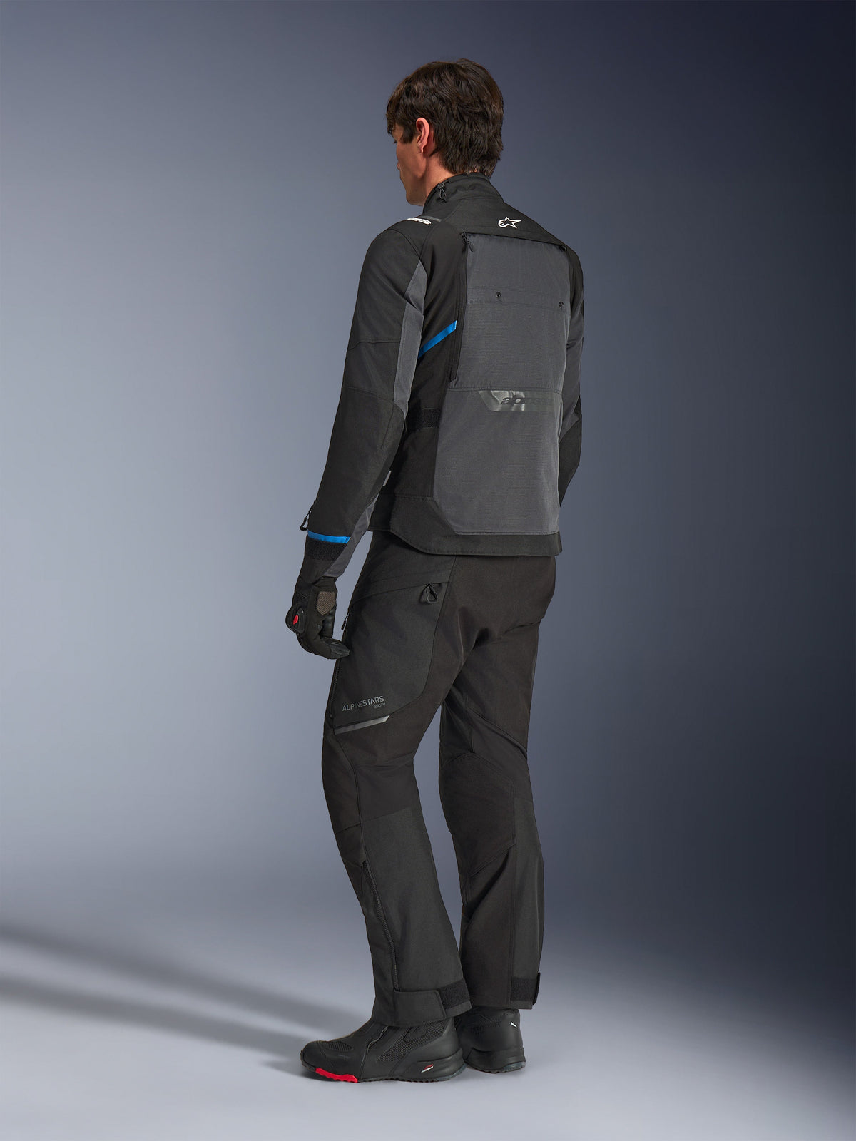 Casaco Alpinestars Cusco Drystar®, Preto e Cinzento Escuro, vista traseira de três quartos mostrando o grande painel de ventilação traseiro e detalhes azuis nas mangas, usado com calças e botas de motociclista pretas para um conjunto de touring completo.