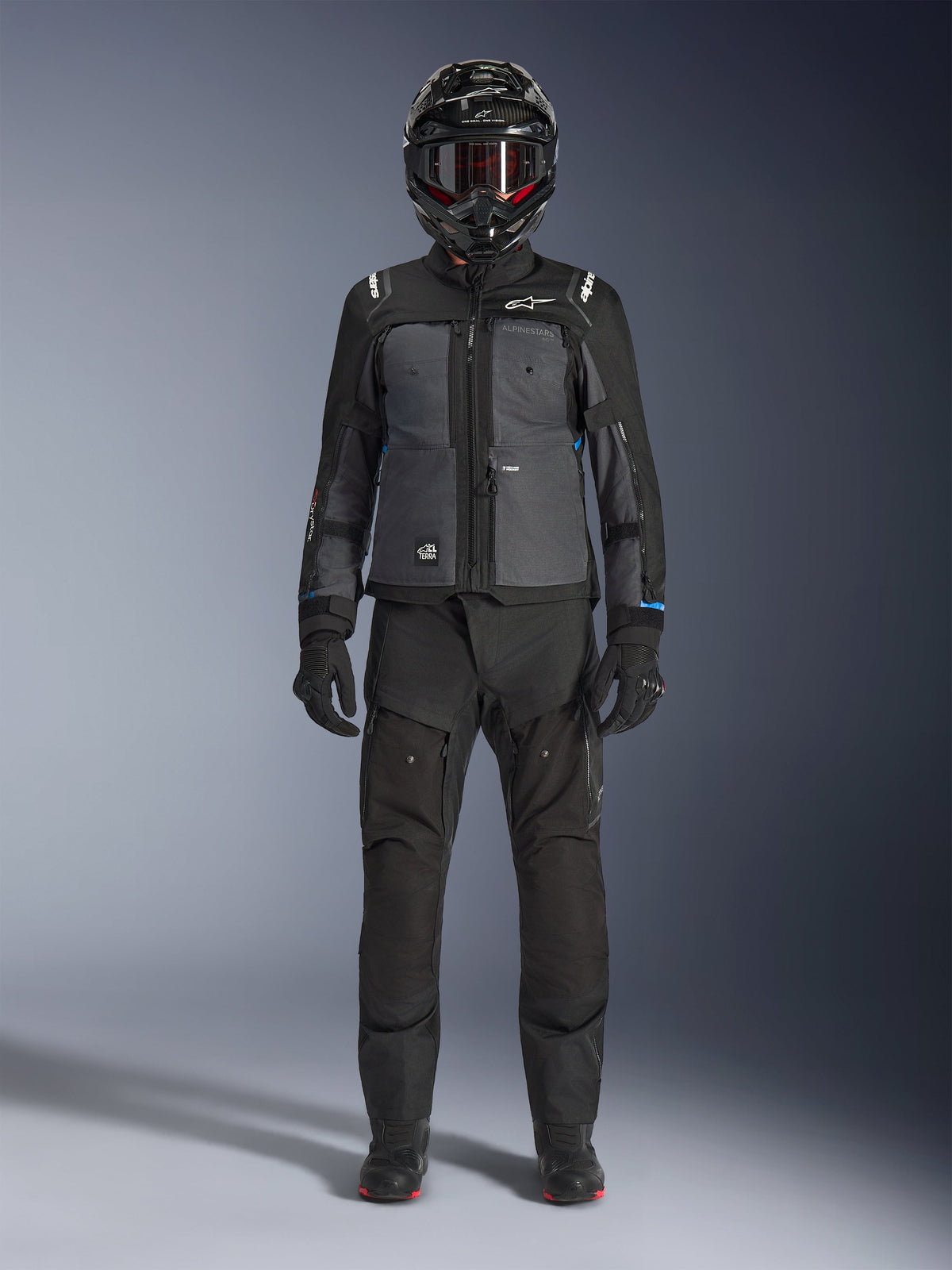 Casaco Alpinestars Cusco Drystar®, casaco de turismo de aventura preto e cinzento escuro, usado com calças pretas a condizer, luvas e um capacete integral, vista frontal de corpo inteiro, vestuário de proteção para motociclismo