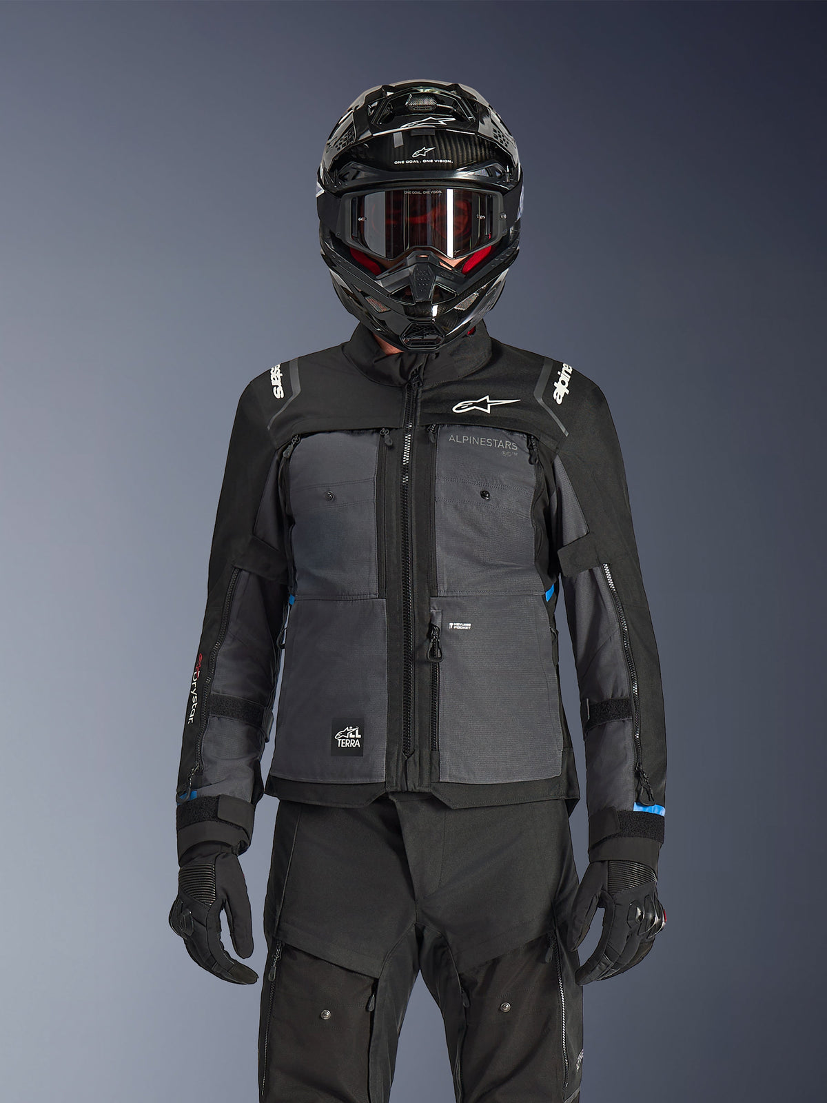 Casaco Alpinestars Cusco Drystar®, preto e cinzento escuro, casaco de motociclismo adventure touring usado com calças técnicas pretas a condizer e um capacete dual-sport, apresentando um design bicolor com painéis em ripstop e logótipos Alpinestars