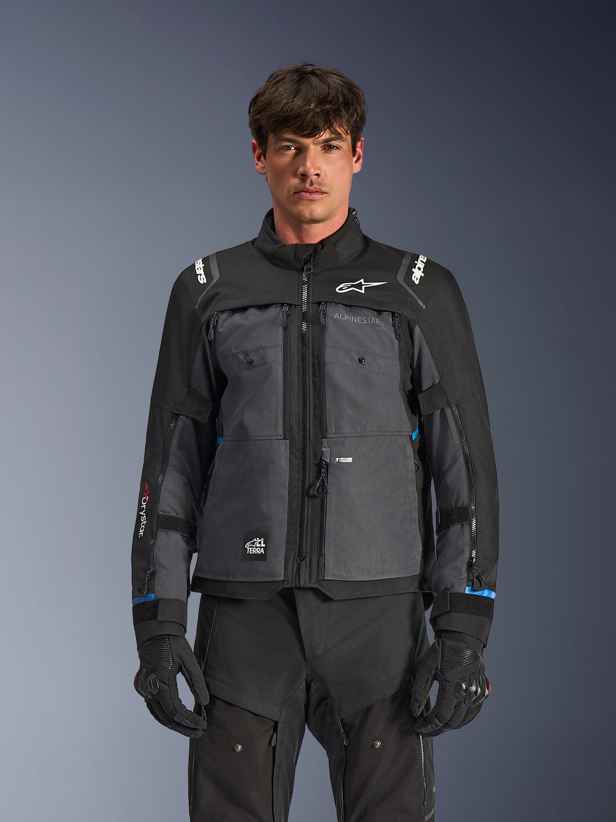 Casaco Alpinestars Cusco Drystar®, casaco de motociclismo touring, preto e cinzento escuro, vista frontal num modelo, com logótipos Alpinestars no peito e ombros, marca Drystar® na manga, design técnico de vários bolsos com fechos de ventilação, combinado com equipamento de condução preto