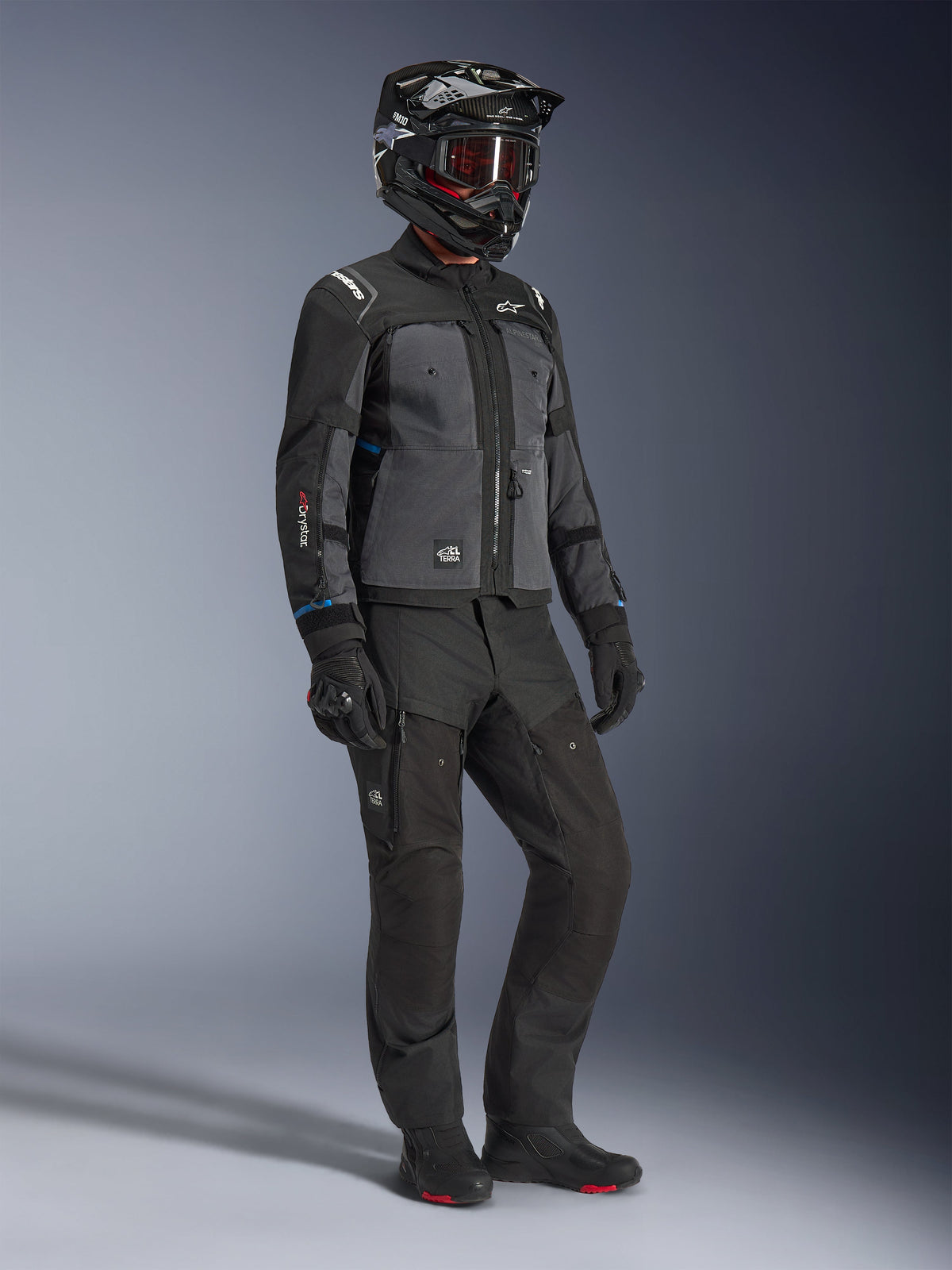 Modelo a usar o Casaco Alpinestars Cusco Drystar® em Preto e Cinzento Escuro, combinado com equipamento completo de motociclismo, incluindo capacete preto, calças técnicas a condizer, luvas e botas, com construção em ripstop e softshell para estrada e turismo de aventura