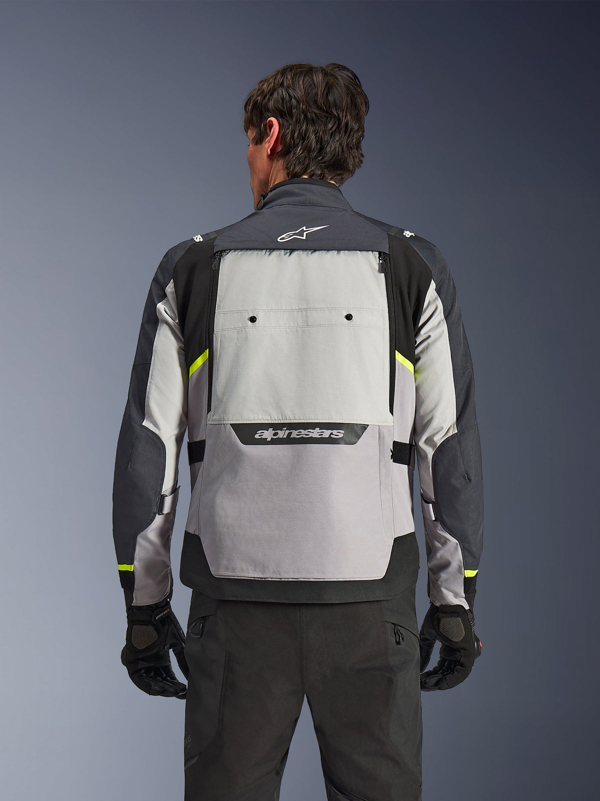 Vista traseira de um motociclista a usar o Casaco Alpinestars Cusco Drystar® em Ice Gray, Mid Gray e Deep Gray, com detalhes em amarelo néon, um grande painel de ventilação traseiro e logótipo Alpinestars em destaque, combinado com calças e luvas de condução pretas.