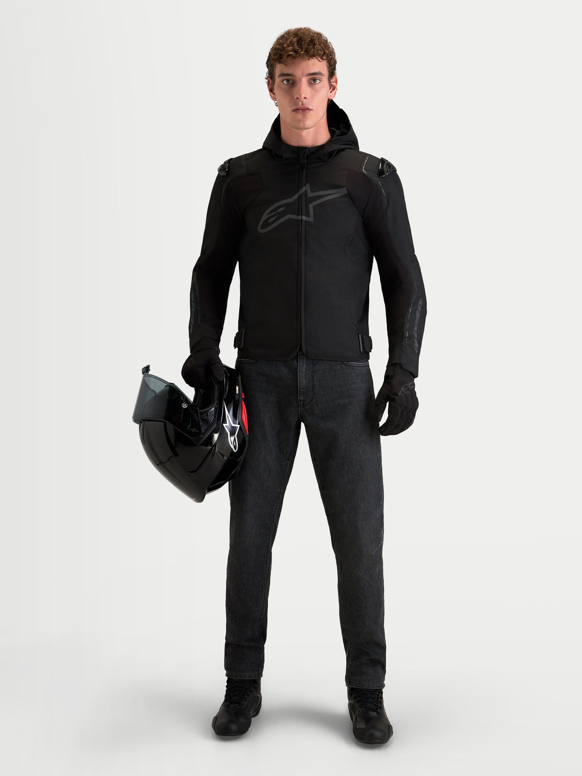 Alpinestars Zaca V2 Waterproof Casaco, casaco de moto em tecido para homem com capuz e proteção nos ombros, preto, modelo a segurar um capacete.