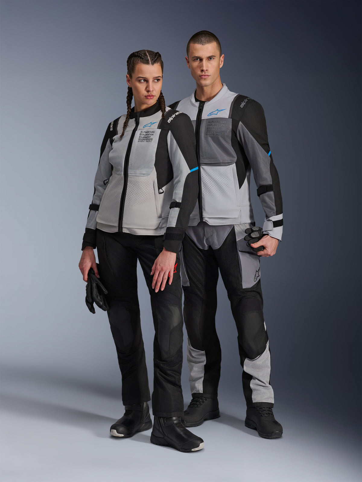 Homem e mulher com casacos Alpinestars Stella ST-2 Air e calças técnicas a condizer, cor Cinzento Gelo e Preto com detalhes azuis, ventilação em malha e marca 'ALPINESTARS EXPLORATION EQUIPMENT', fotografia de estúdio.