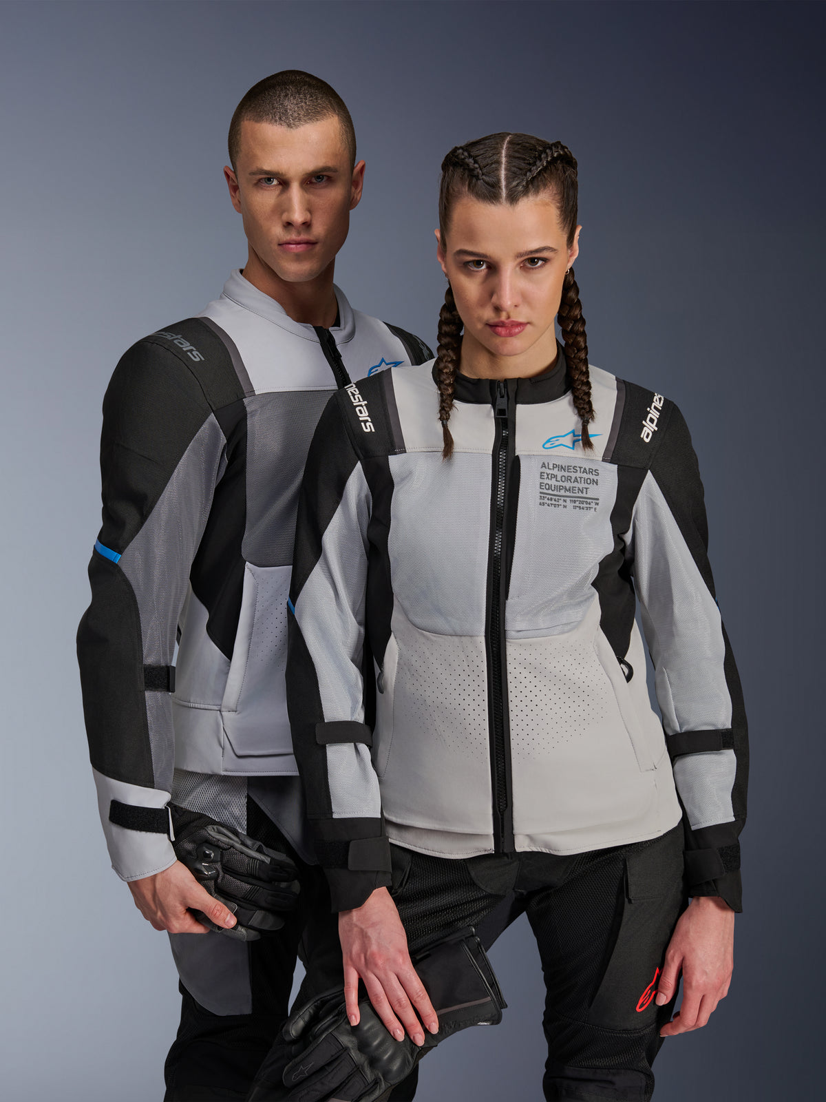 Casacos Alpinestars ST-2 Air e Stella ST-2 Air, Cinza Gelo e Preto, casacos de motociclismo em têxtil com painéis em malha e perfurações a laser, com a marca 'Alpinestars Exploration Equipment', usados por modelos masculino e feminino a segurar luvas de motociclismo