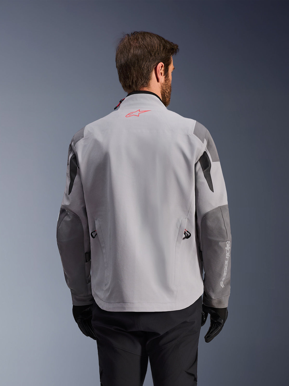 Alpinestars Maxdura Drystar®XF Laminated Armored Shell, vista traseira de casaco de motociclismo de aventura, em cinzento médio e cinzento escuro, com um logótipo de estrela vermelha na gola e marca vertical branca na manga, usado com luvas e calças de condução pretas