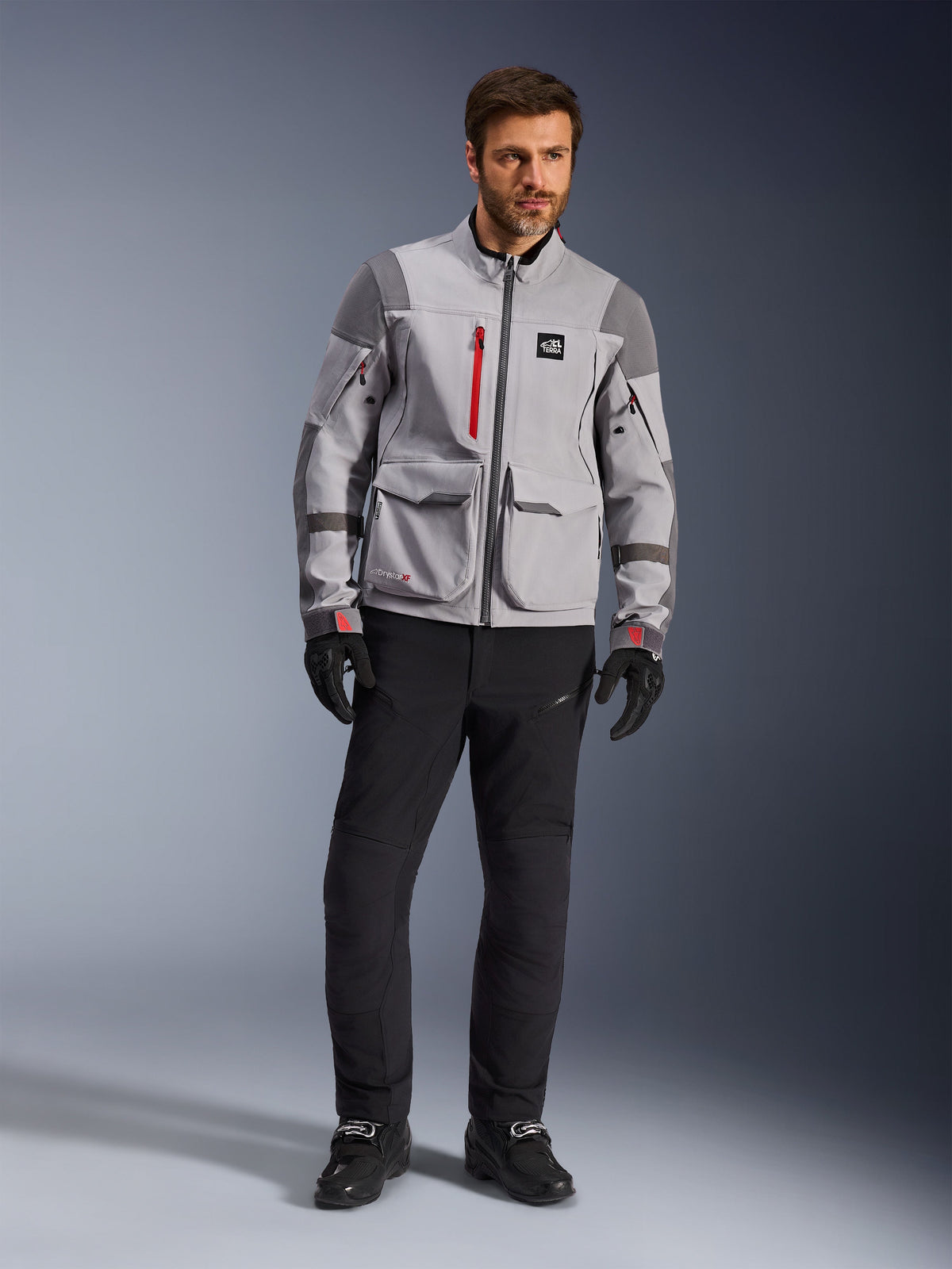 Alpinestars Maxdura Drystar®XF Laminated Armored Shell, casaco de motociclismo de aventura, cinzento médio e cinzento escuro com pormenores vermelhos, usado com calças de condução pretas e luvas, vista frontal realçando a construção técnica e os grandes bolsos cargo.