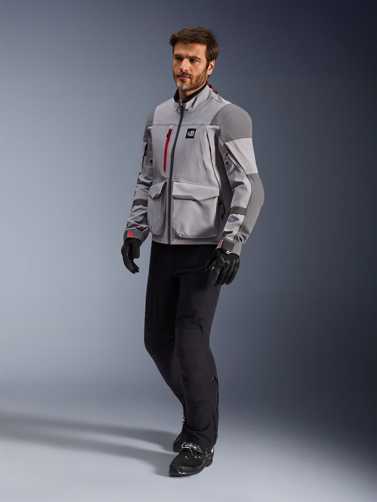 Alpinestars Maxdura Drystar®XF Laminated Armored Shell, casaco de motociclismo de aventura, cinzento médio e cinzento escuro, vestido por um modelo com calças de condução e luvas pretas, com detalhes de fechos vermelhos e bolsos cargo, equipamento técnico profissional para todas as condições meteorológicas para touring de aventura