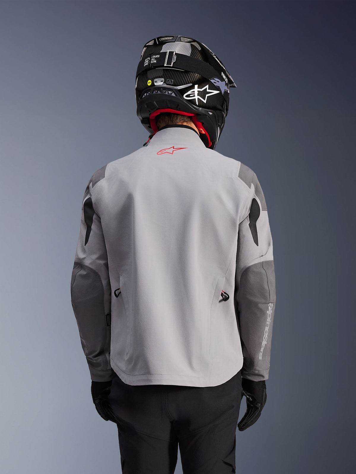Alpinestars Maxdura Drystar®XF Laminated Armored Shell, casaco de motociclismo de aventura, cinzento médio e cinzento escuro, vista traseira mostrando o corpo cinzento claro com painéis reforçados em cinzento mais escuro nos ombros e cotovelos, logótipo Alpinestars vermelho na parte superior das costas, usado com capacete preto e luvas pretas.