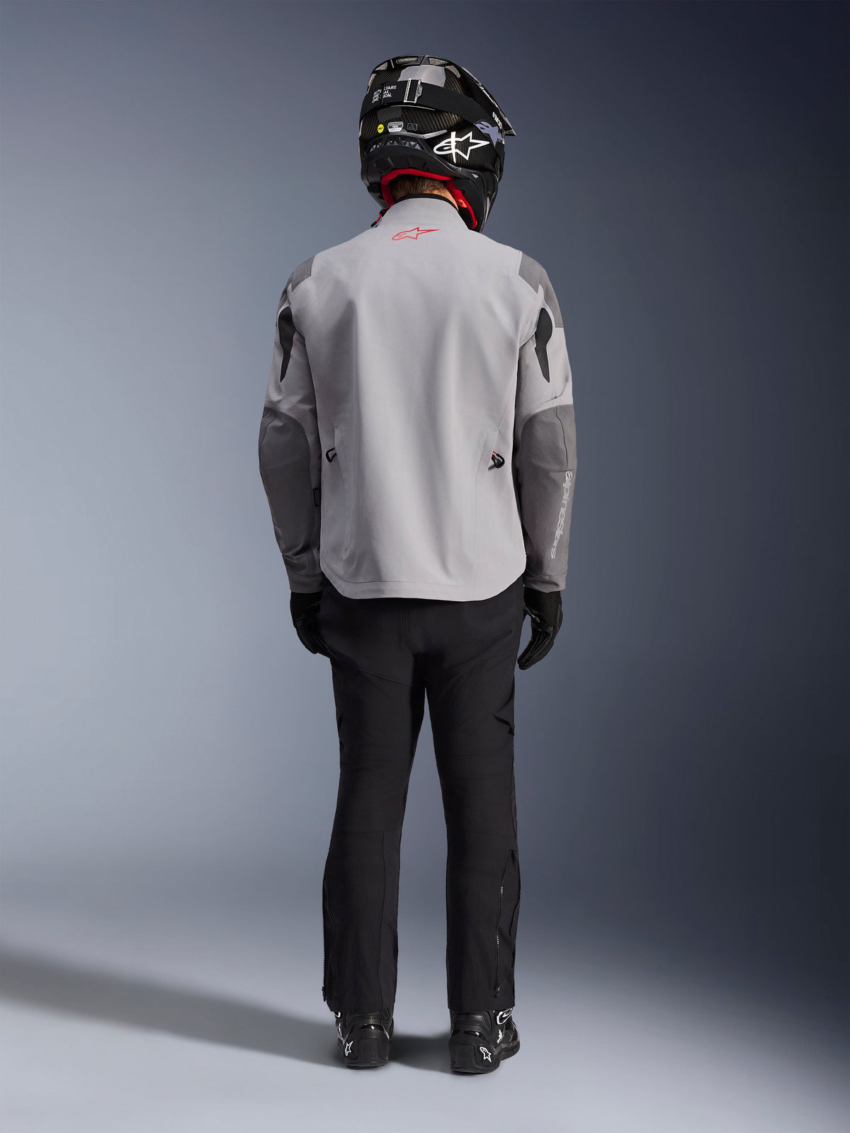 Alpinestars Maxdura Drystar®XF Laminated Armored Shell, casaco de motociclista em Cinzento Médio e Cinzento Escuro, visto de costas num motociclista, usado com calças pretas, botas pretas e um capacete preto e branco, equipamento de touring de aventura