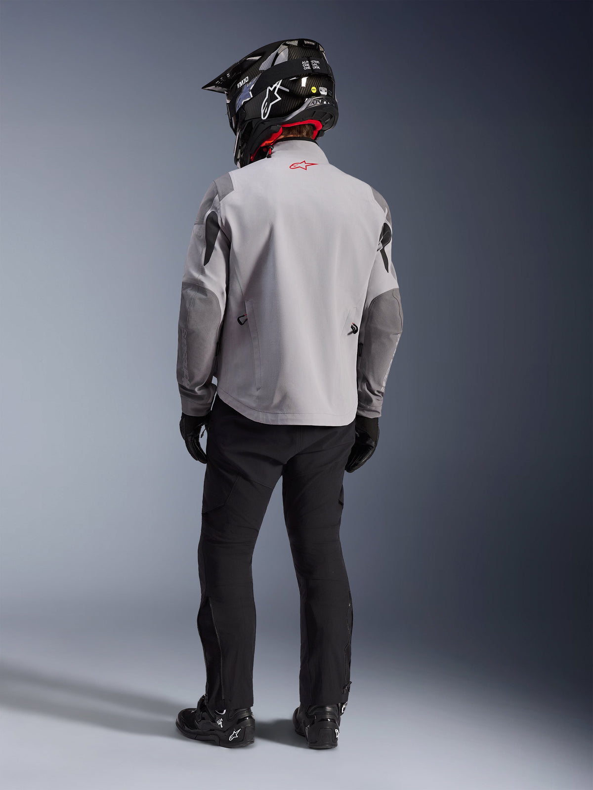 Vista traseira de um motociclista a usar o casaco Alpinestars Maxdura Drystar®XF Laminated Armored Shell em cinzento médio e cinzento escuro, com um logótipo de estrela vermelha na gola, combinado com calças de aventura pretas, um capacete preto com padrão de carbono e botas pretas.