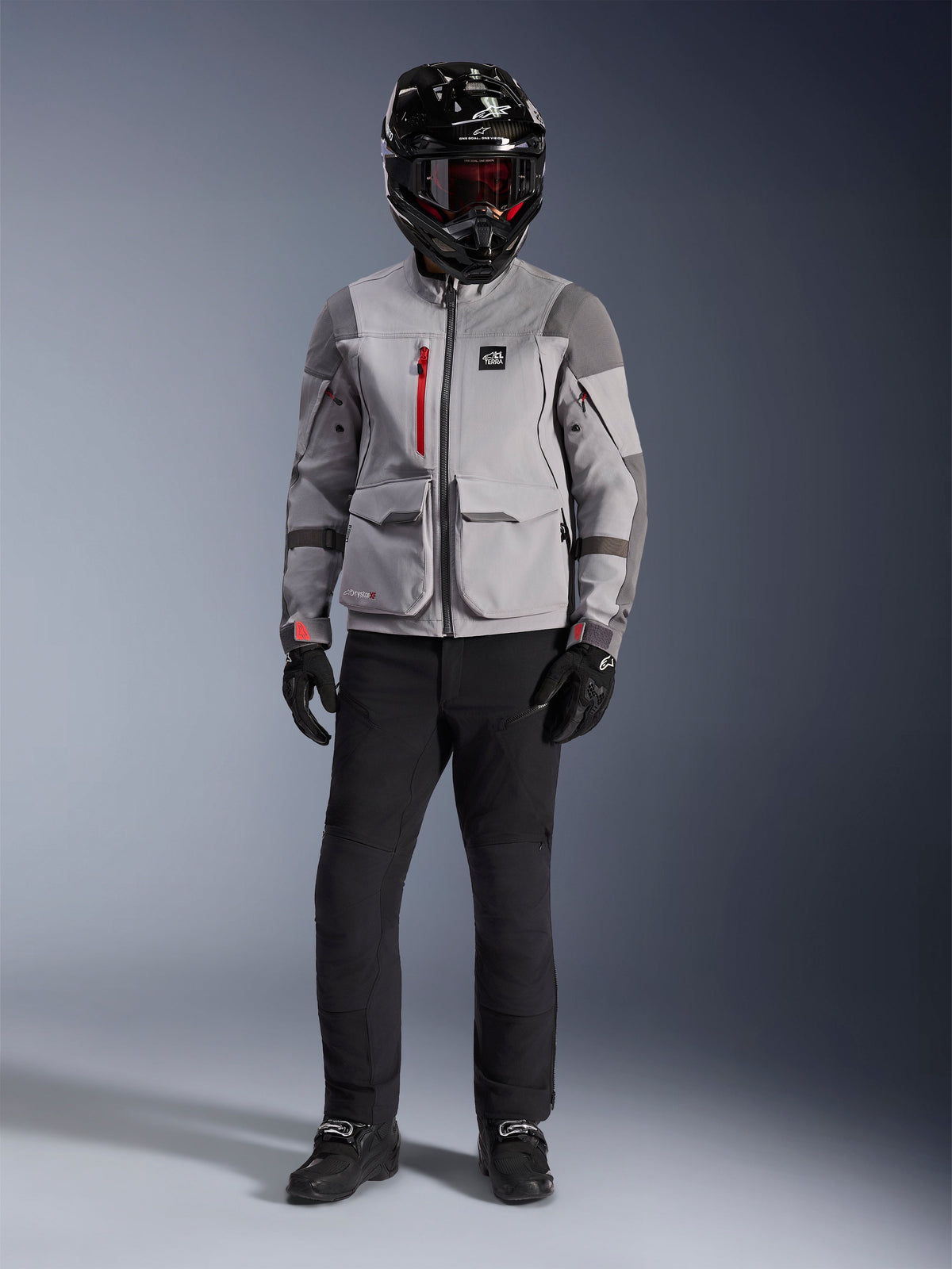 Alpinestars Maxdura Drystar®XF Laminated Armored Shell, casaco de aventura cinzento médio e cinzento escuro com detalhes de fechos vermelhos, usado por um motociclista com capacete preto, óculos vermelhos e calças pretas, equipamento de motociclismo profissional para todas as condições meteorológicas