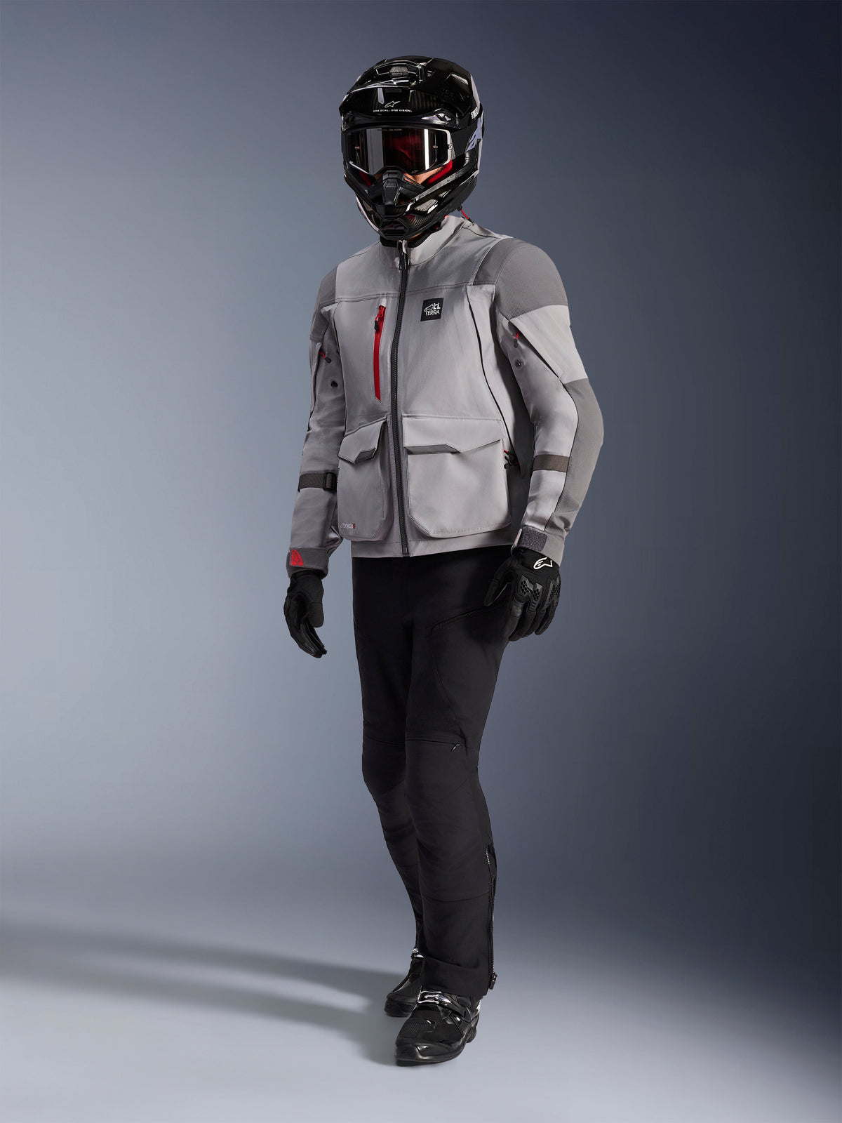 Motociclista de aventura a usar o casaco Alpinestars Maxdura Drystar®XF Laminated Armored Shell, casaco de motociclismo em cinzento médio e cinzento escuro, capacete de aventura preto com óculos, calças e luvas pretas, com detalhes de fechos vermelhos e bolsos cargo, equipamento técnico para adventure touring.