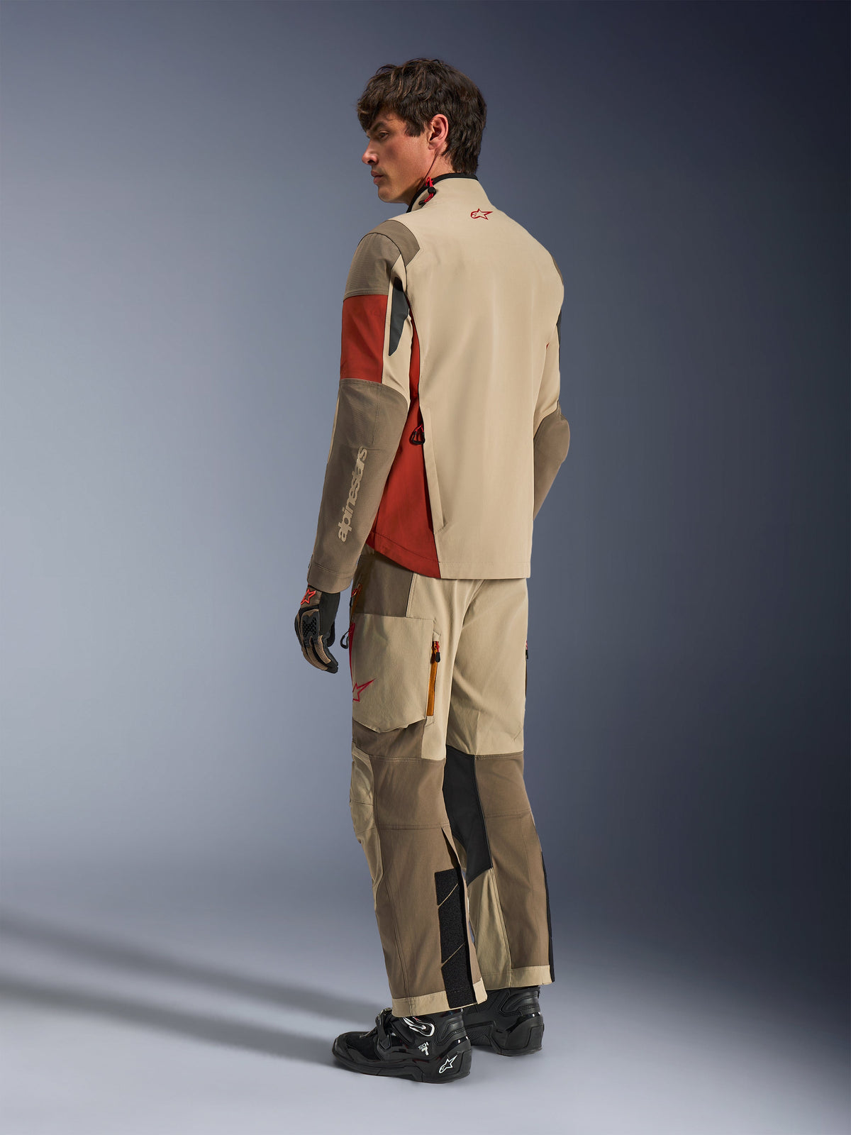 Modelo masculino a usar a Alpinestars Maxdura Drystar®XF Laminated Armored Shell em Aluminum Walnut Rust Brown, calças de adventure touring a condizer, luvas pretas e botas pretas, vista traseira que mostra a construção têxtil técnica e as zonas reforçadas.