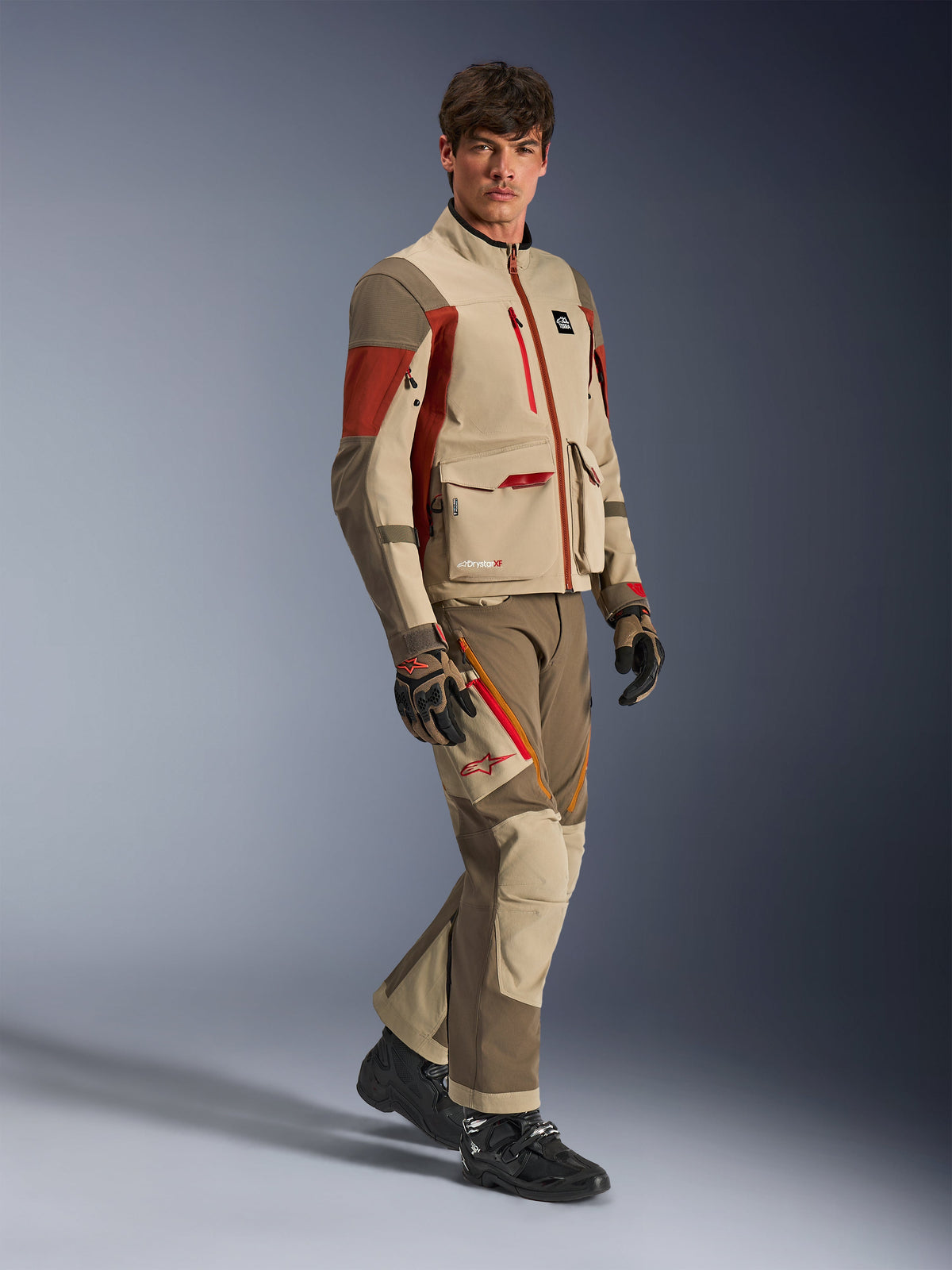 Modelo masculino a usar o casaco de aventura Alpinestars Maxdura Drystar®XF Laminated Armored Shell na cor Aluminum Walnut Rust Brown com calças a condizer, luvas pretas e castanhas e botas pretas, apresentando um design técnico multicamada com bolsos de carga e detalhes de fechos vermelhos, num conjunto coordenado para adventure touring
