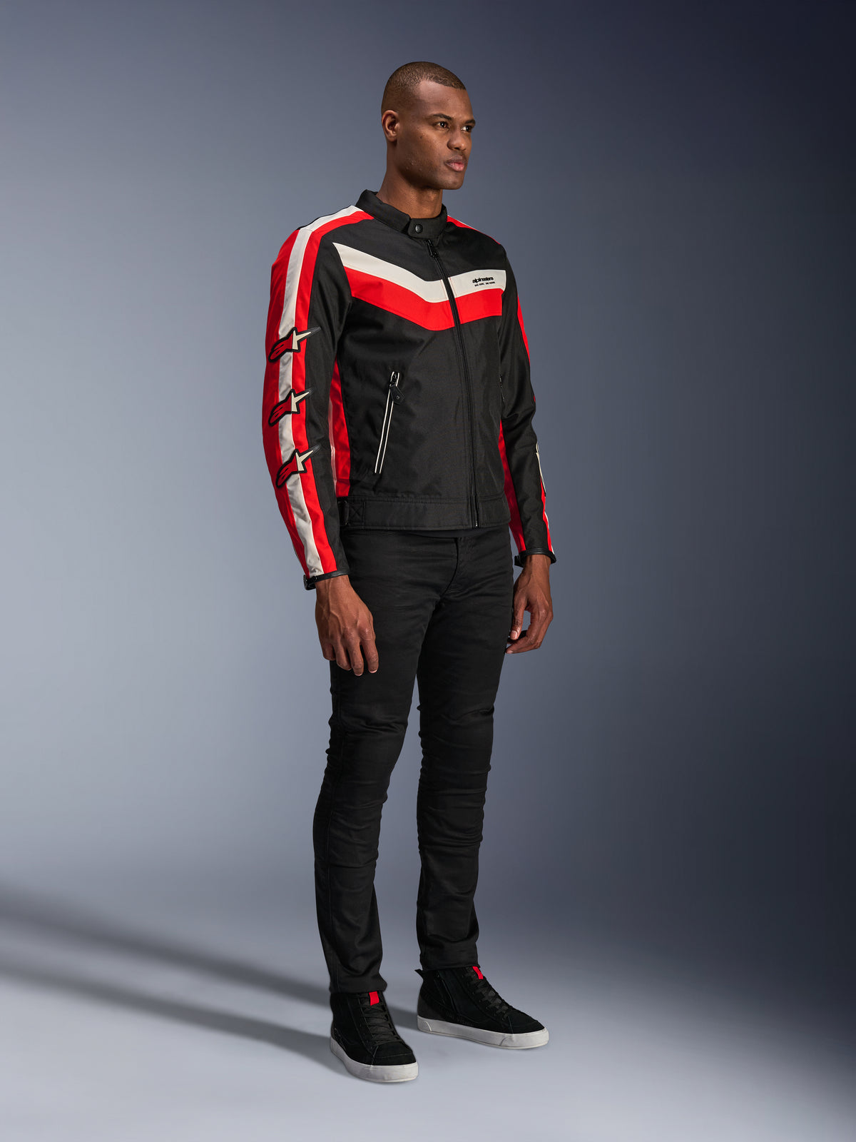 Modelo masculino a usar o Casaco Alpinestars T-Dyno WR, casaco de motociclismo têxtil em Black Bright Red Ecru, com riscas vermelhas e ecru e logótipos de estrela Alpinestars nas mangas, combinado com calças pretas e sapatilhas de cano alto pretas, equipamento de condução urbana de inspiração vintage.