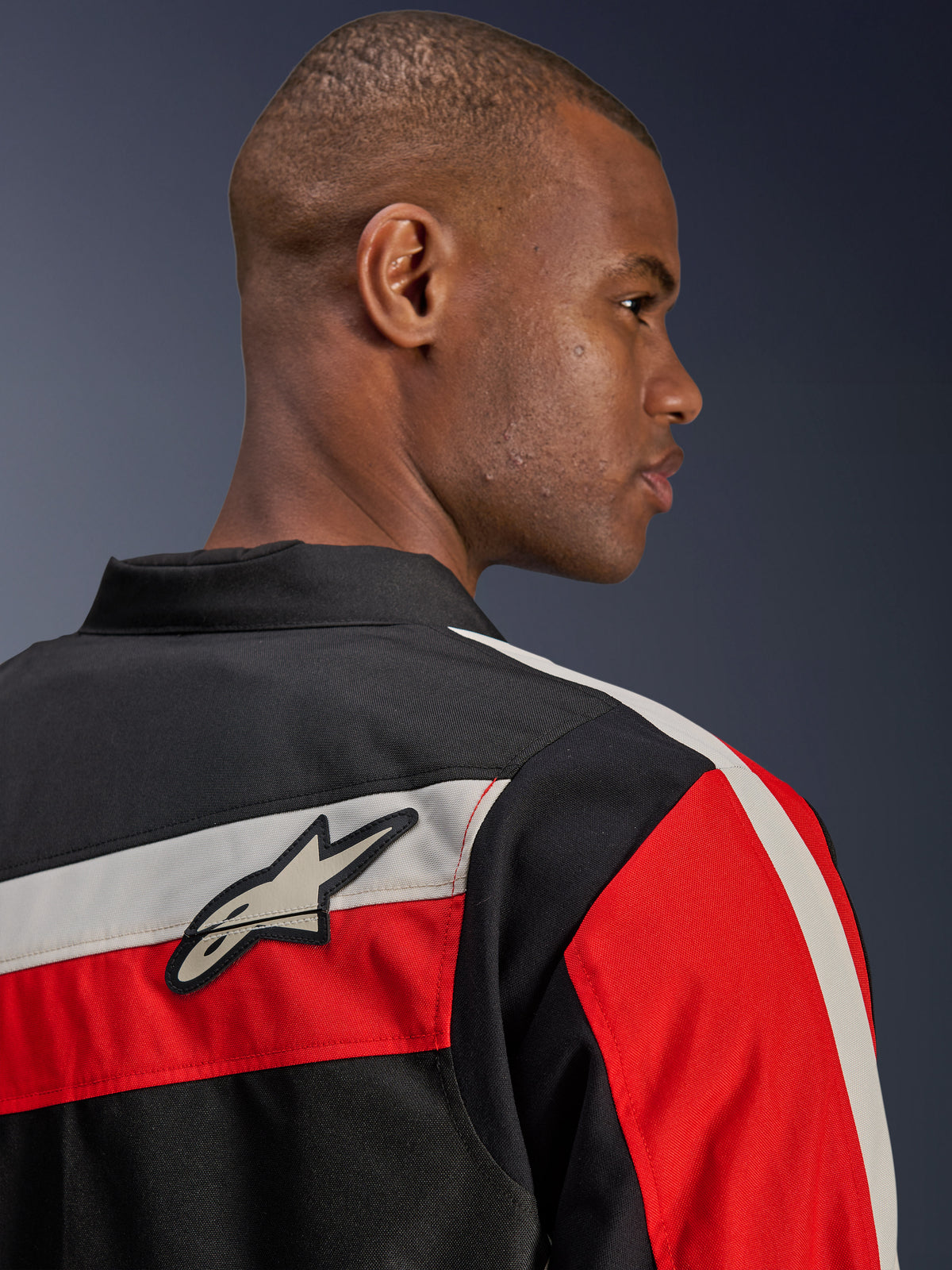 Casaco Alpinestars T-Dyno WR, vista aproximada das costas num modelo, casaco de motociclismo têxtil em preto com riscas horizontais cru e vermelho vivo, logótipo de estrela da Alpinestars no ombro, estilo de motociclista de inspiração vintage