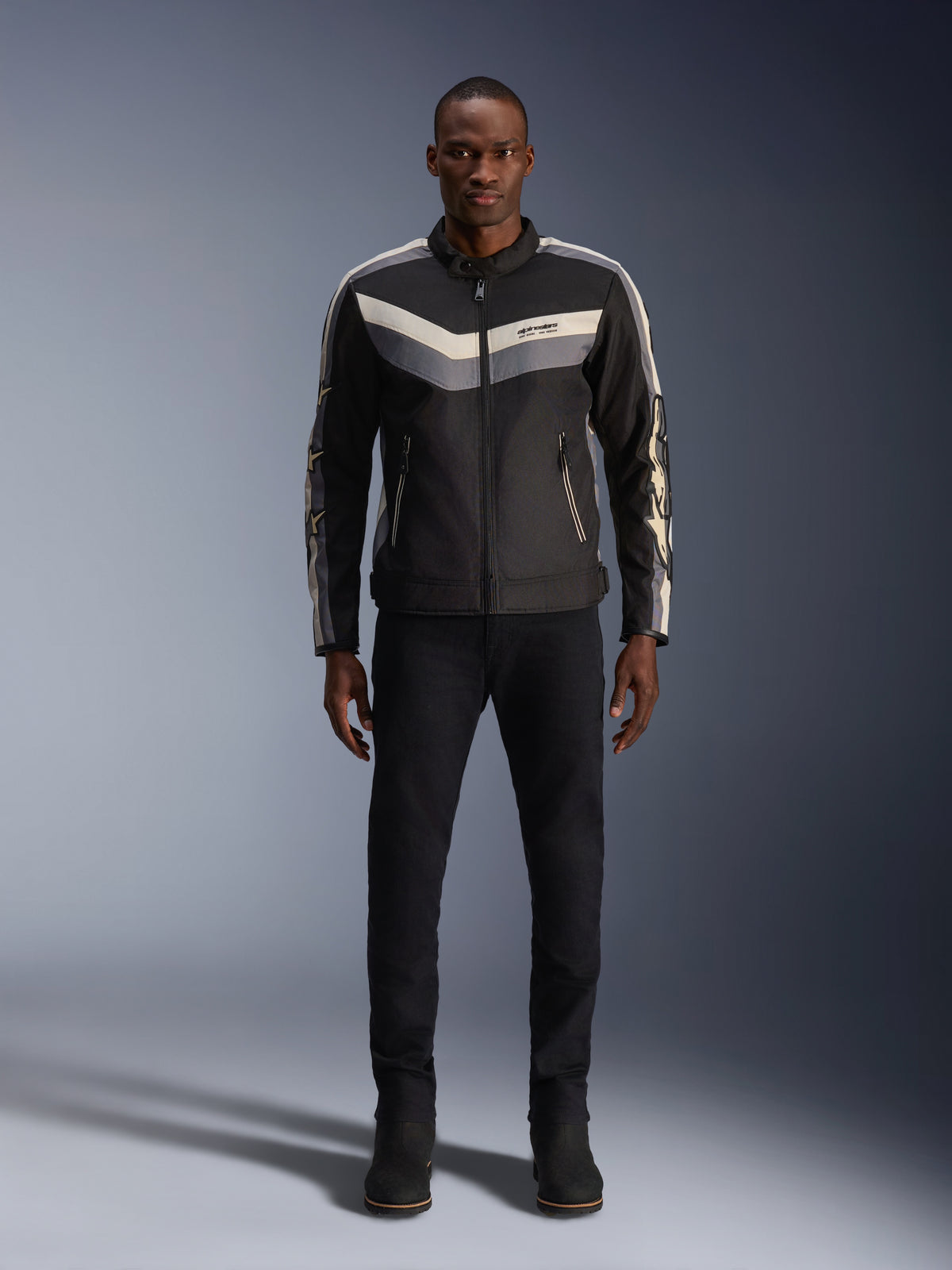 Casaco Alpinestars T-Dyno WR, preto, cinzento escuro e ecru, casaco de motociclismo têxtil com padrão chevron no peito de inspiração vintage, usado por modelo masculino com calças de condução e botas pretas, equipamento de estrada urbano contemporâneo