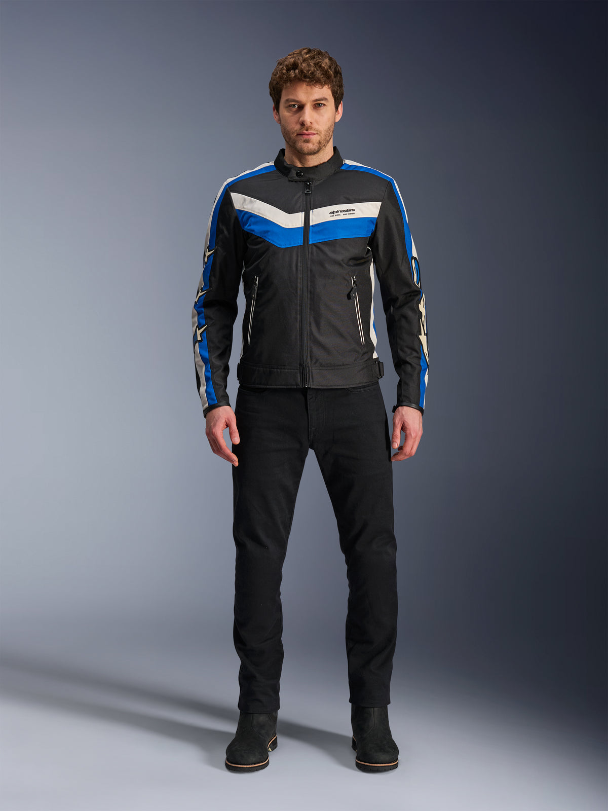 Casaco Alpinestars T-Dyno WR, casaco de motociclismo têxtil, preto, azul e cru, usado por modelo masculino com calças e botas pretas, apresentando riscas em chevron no peito e design de herança motociclista