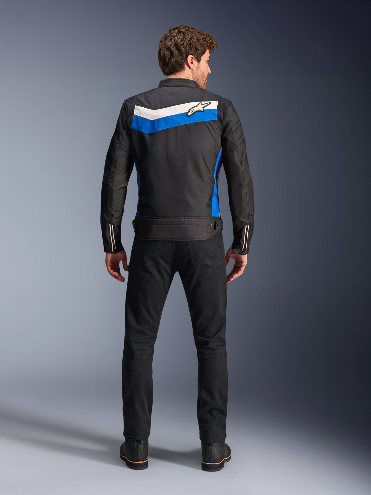 Alpinestars T-Dyno WR Jacket, casaco de motociclismo têxtil, cor Preto Azul Ecru, vista traseira completa em modelo, com uma risca em chevron azul e off-white com o logótipo Alpinestars, combinado com calças e botas pretas, equipamento de condução urbana de inspiração vintage
