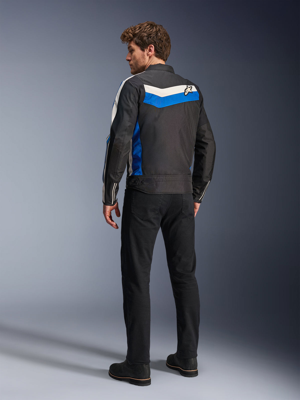 Casaco Alpinestars T-Dyno WR em Preto Azul Ecru, vista traseira num modelo masculino, com uma risca em chevron branca e azul nos ombros e logótipo de estrela, casaco de motociclismo têxtil de inspiração vintage, usado com calças e botas pretas