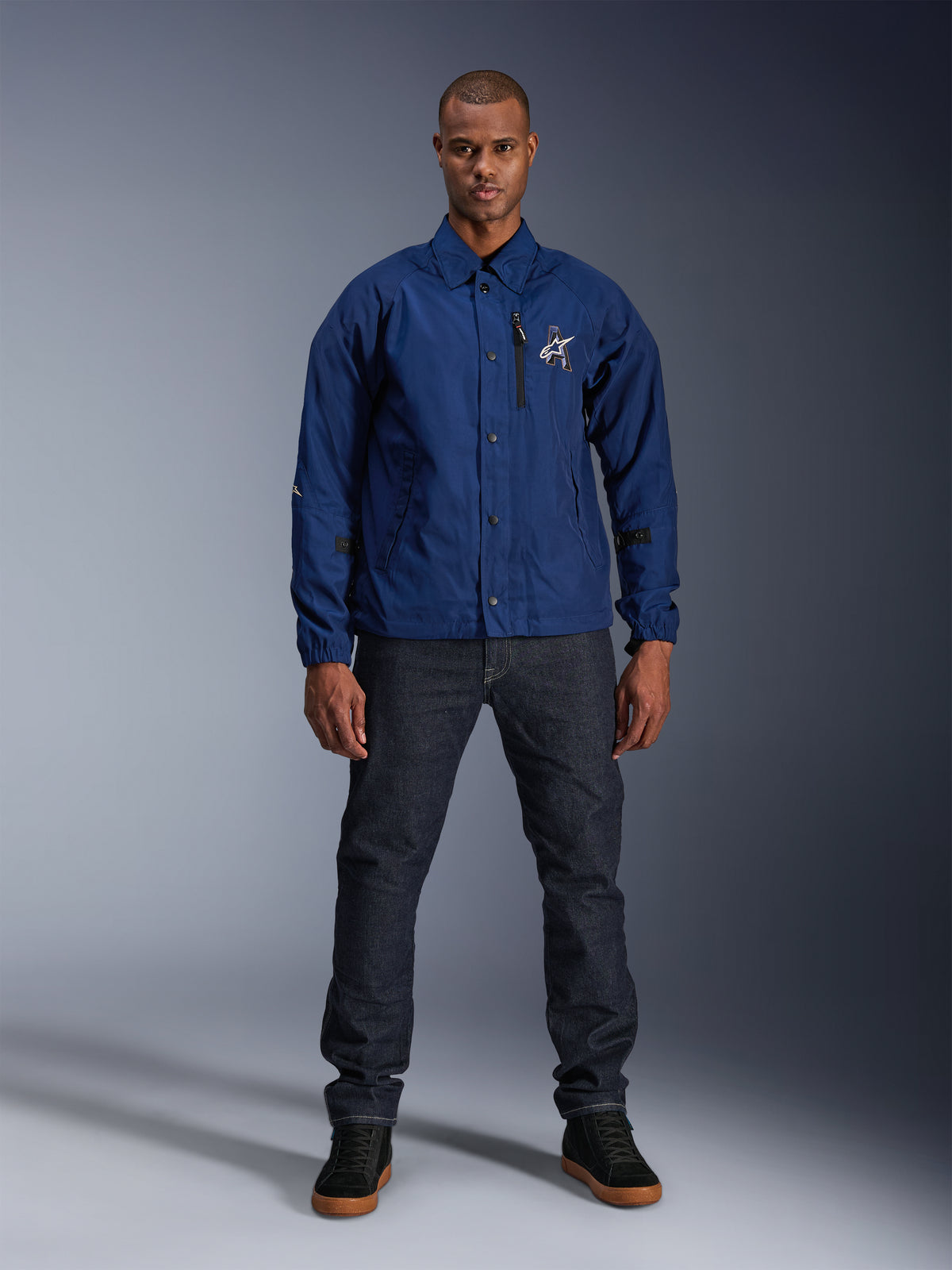 Casaco Alpinestars Revved WR, azul-marinho, casaco de motociclismo estilo 'coach', usado por um homem com calças de ganga escuras e ténis pretos, com fecho de botões de pressão e logótipo Alpinestars em 3D, streetwear urbano contemporâneo