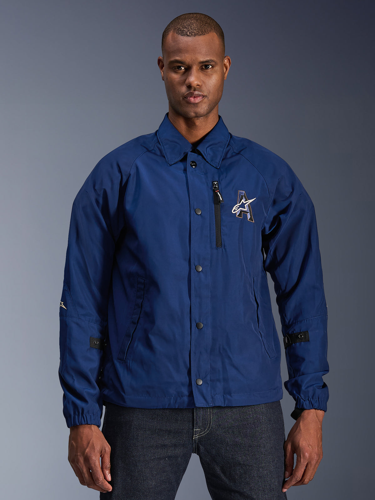 Alpinestars Revved WR Jacket, casaco de motociclismo urbano para homem, azul escuro, estilo coach com gola dobrável e botões de pressão frontais, bolso no peito com fecho de correr preto, logótipo grande bordado em 3D, tiras ajustáveis nas mangas, material resistente à água com acabamento acetinado