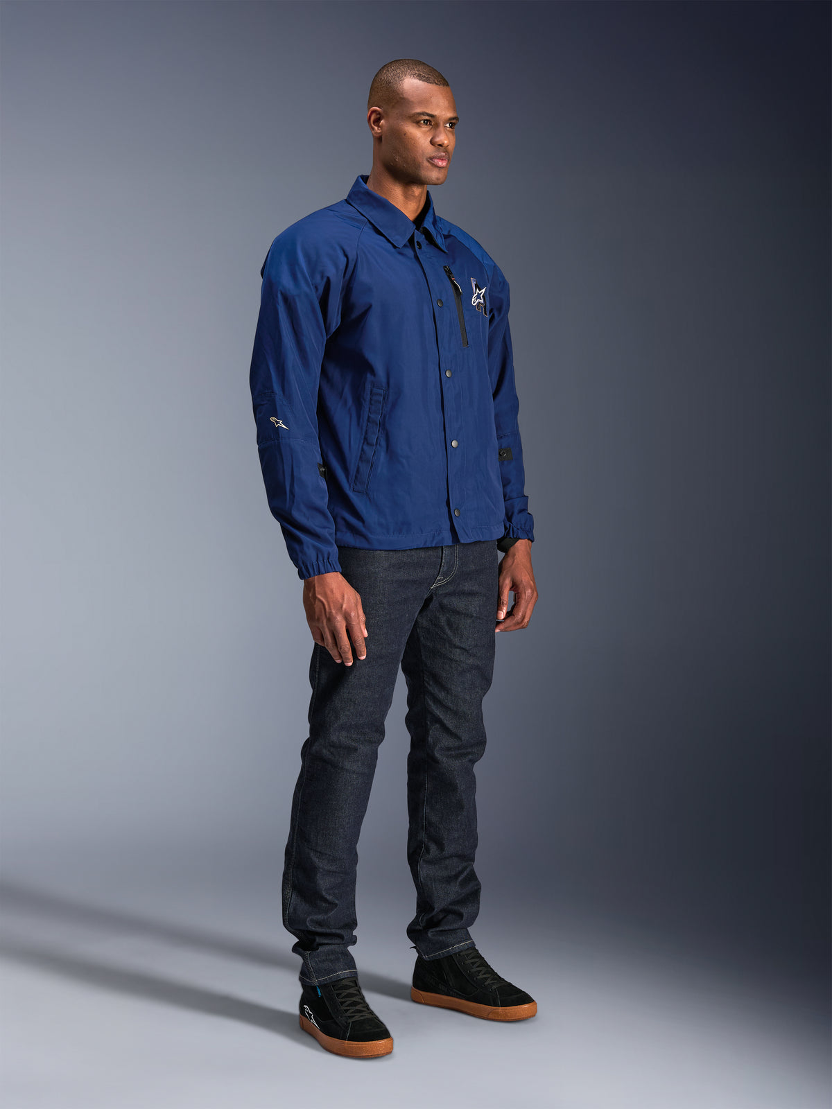 Casaco Alpinestars Revved WR, casaco de motociclismo azul, usado por um modelo masculino com calças de ganga escuras e ténis pretos, com uma silhueta de casaco de treinador resistente à água, botões de pressão frontais e logótipo minimalista no peito